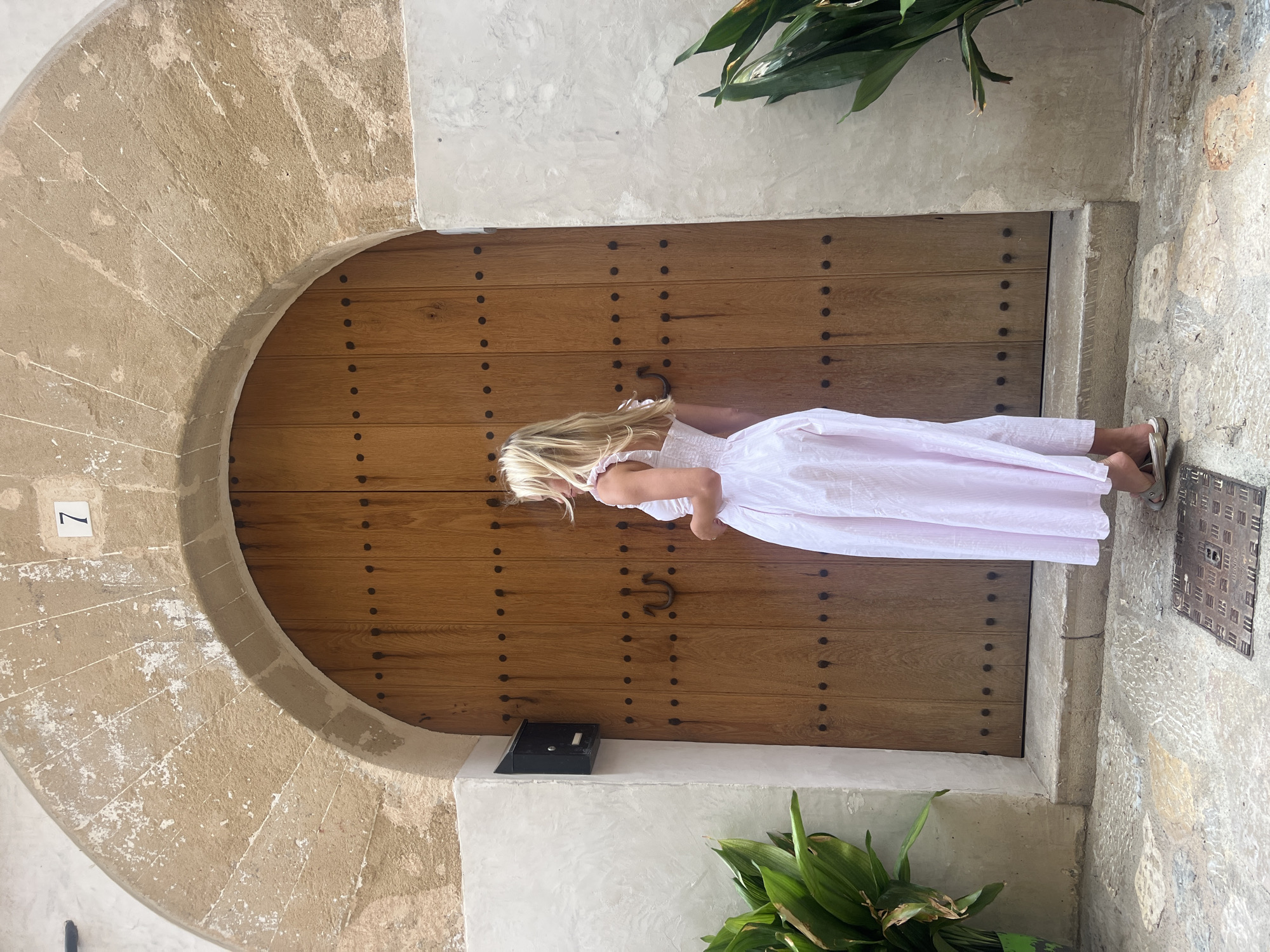 A woman in a pink sundress stands with her back to the camera, facing a large, arched wooden door in Pollença, Spain. The door is made of vertical planks and is studded with dark metal rivets. A dark metal horseshoe-shaped door pull is visible in the center. Above the door, a stone archway is adorned with peeling white paint and a small, white plaque with the number "7". To the left of the door, a black mailbox is mounted on a rough, textured wall. Lush green plants flank the entrance, with one on the left in a pot and another on the right growing from the ground. The ground in front of the door is paved with irregular stone tiles and a metal grate is visible in the foreground. The overall scene suggests an old, rustic building in a Mediterranean town.