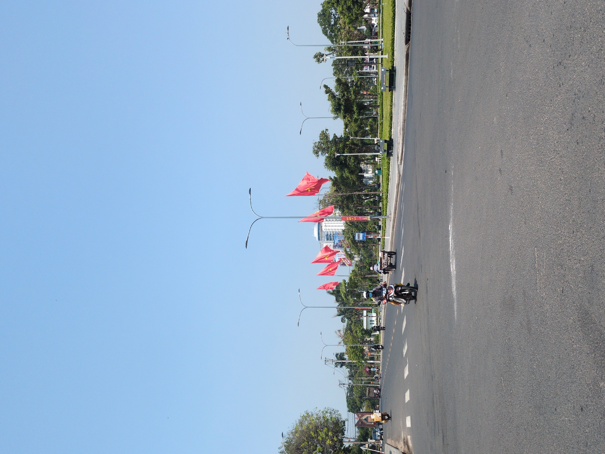The image captures a sunny day on a wide avenue in Tuy Hòa, Vietnam. The view is along the street, showing the road stretching into the distance. Red Vietnamese flags are displayed along the median strip, attached to lampposts. Several motorbikes are traveling along the road. The sky is clear and blue. Green trees and buildings are visible alongside the road.