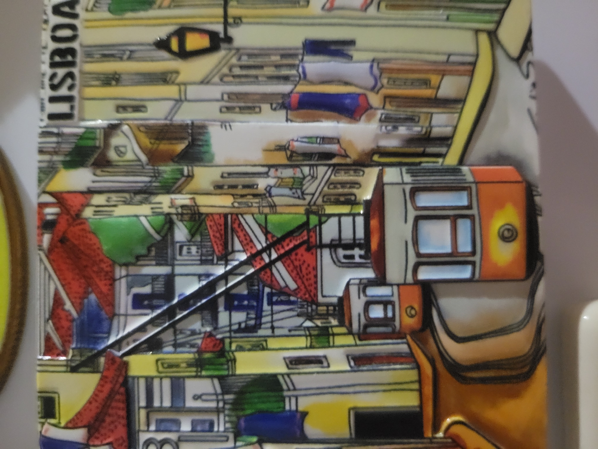 The image depicts a ceramic tile featuring a stylized illustration of a city scene. Two orange and white trams are the prominent subjects, positioned on a curved street with tram tracks. The background is filled with colorful buildings in shades of yellow, red, green, and white, characterized by detailed windows, balconies, and tiled roofs. Laundry hangs from some windows, adding a touch of domestic detail. The word "LISBOA" is visible in black lettering at the top right of the tile, indicating the city of Lisbon, Portugal. The overall style is illustrative and vibrant, with bold outlines and flat areas of color. The scene is not located in Belaazyorsk, Belarus, as this is an illustration of Lisbon.