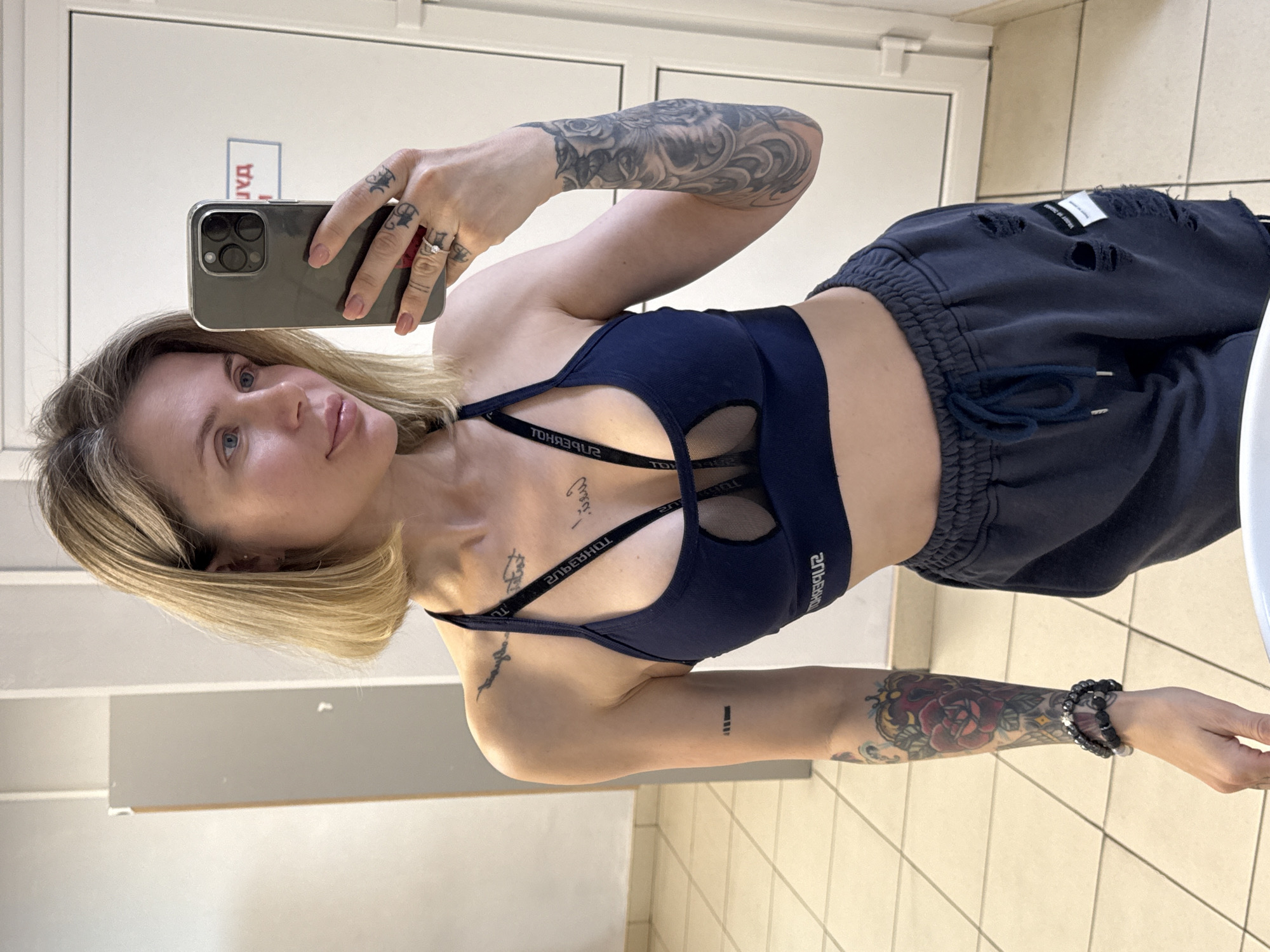 A woman with blonde hair is taking a selfie in a bathroom with a smartphone. She is wearing a dark blue sports bra and dark blue sweatpants with rips. Her left arm is heavily tattooed with various designs, and she is also wearing a beaded bracelet on her left wrist. Additional tattoos are visible on her chest and right arm. The bathroom has white walls and tiled floors. A sign with Cyrillic text is visible on a door in the background. The image was taken in Mayma, Russia.