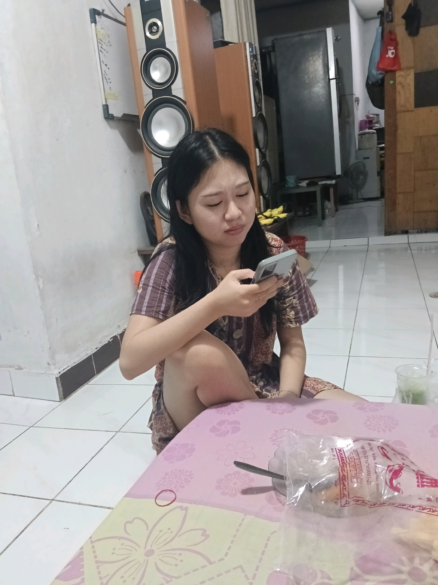 In Jatimulya, Indonesia, a young woman of East Asian descent sits on the tiled floor of a room, engaged with her smartphone. She wears a short-sleeved, patterned dress in shades of brown and purple. Her knees are bent, and her body is angled towards her left. To her right, a pink and green patterned fabric covers a surface, possibly a table, adorned with floral designs. On this fabric, a metal spoon rests inside a silver bowl, which is partially obscured by a clear plastic bag containing food items. The room's background features a tall, dark refrigerator, a large speaker system stacked vertically, and wooden cabinetry. The lighting suggests an indoor setting, likely residential.