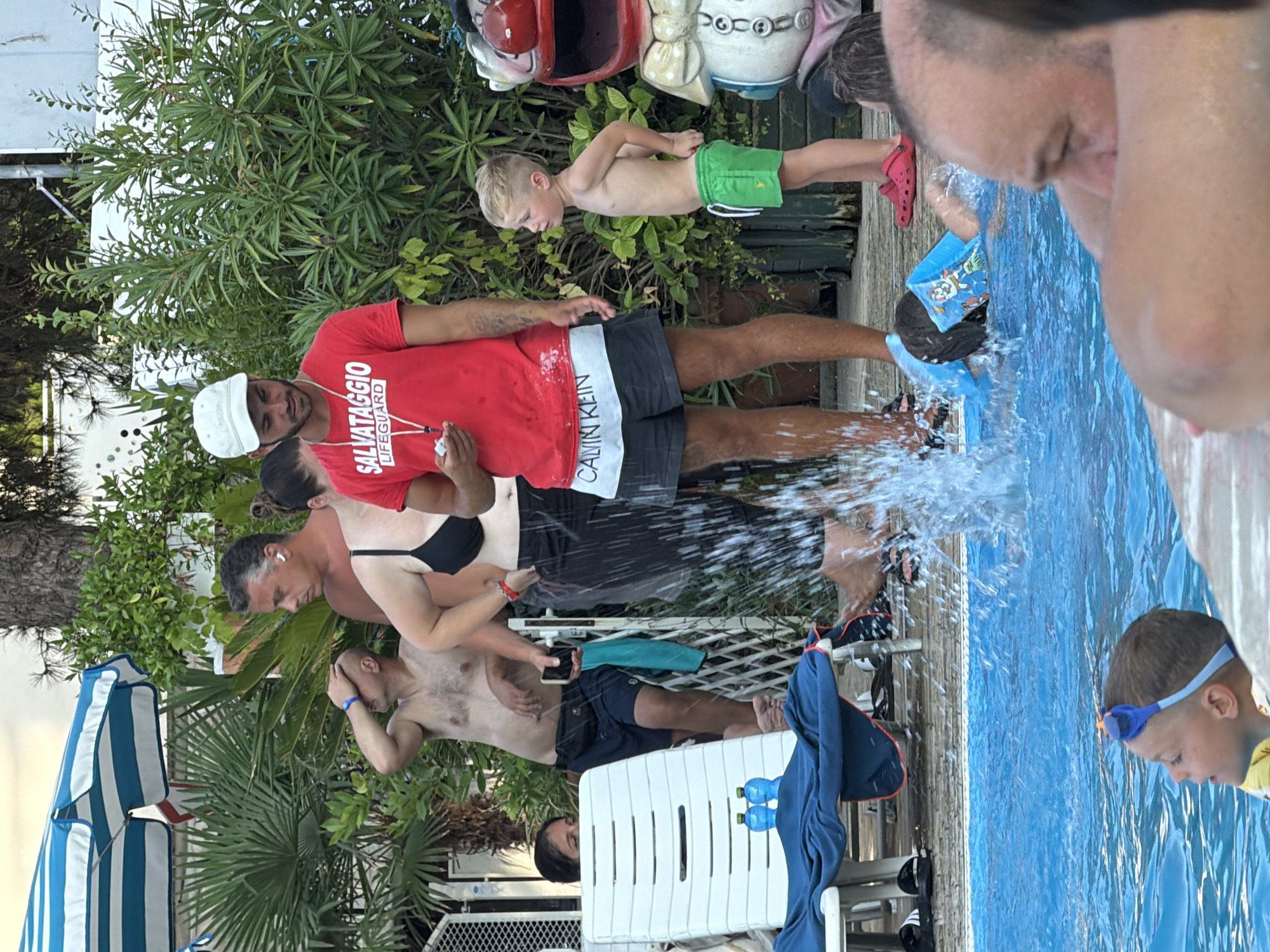 This media file shows a group of people near a swimming pool on a sunny day, likely in Gatteo, Italy. The image is oriented vertically, with the sky at the bottom and the ground at the top.
In the foreground, a young boy wearing blue swimming goggles looks down into the pool, his face partially obscured by the water's reflection. He appears to be in or near the water. To his left, a man in a red t-shirt with "SALVATAGGIO LIFEGUARD" printed on it is partially visible. He is holding a phone and seems to be interacting with others.
A woman in a black bikini top is standing next to him, embracing a man. Behind them, a young boy in green shorts stands near a wooden deck, looking away from the camera. Other individuals and parts of their bodies are visible, some in the water and some on the poolside. There are lounge chairs and greenery surrounding the pool area.
The activity taking place appears to be a leisure day at a swimming pool, possibly with some supervision given the "LIFEGUARD" shirt. The mood seems relaxed and familial, with people enjoying the water and the outdoors. The bright sunlight and clear blue water suggest good weather. The presence of lounge chairs and the pool itself indicates a resort or recreational area. The text "SALVATAGGIO LIFEGUARD" and "CALVIN KLEIN" are visible on clothing.