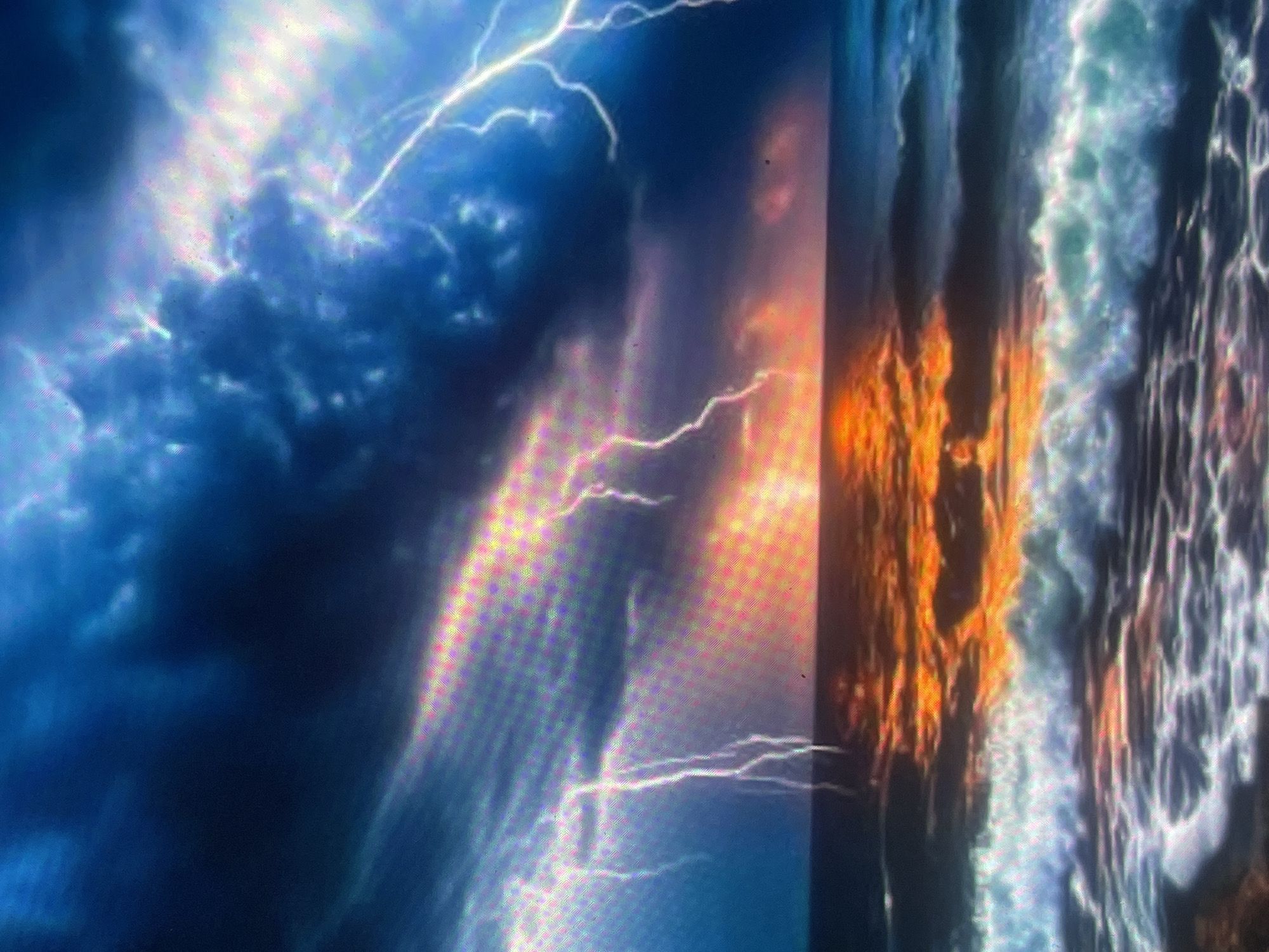 The image depicts a dramatic seascape under a violent electrical storm, located in the city of Zelenyi Hai, Ukraine. The sky is dominated by dense, dark storm clouds in deep blue and grey tones, illuminated by numerous bright white and bluish-white lightning strikes that fork across the upper frame. On the horizon, a vivid orange and red glow, suggestive of either a sunrise or sunset, casts an intense, fiery reflection across the turbulent ocean surface. The sea exhibits choppy waves, with white foam cresting in the foreground and mid-ground, mirroring both the somber stormy sky and the vibrant horizon light. No people, specific man-made objects, or identifiable urban features are visible within the scene. The environment is one of overwhelming natural power and contrast.
