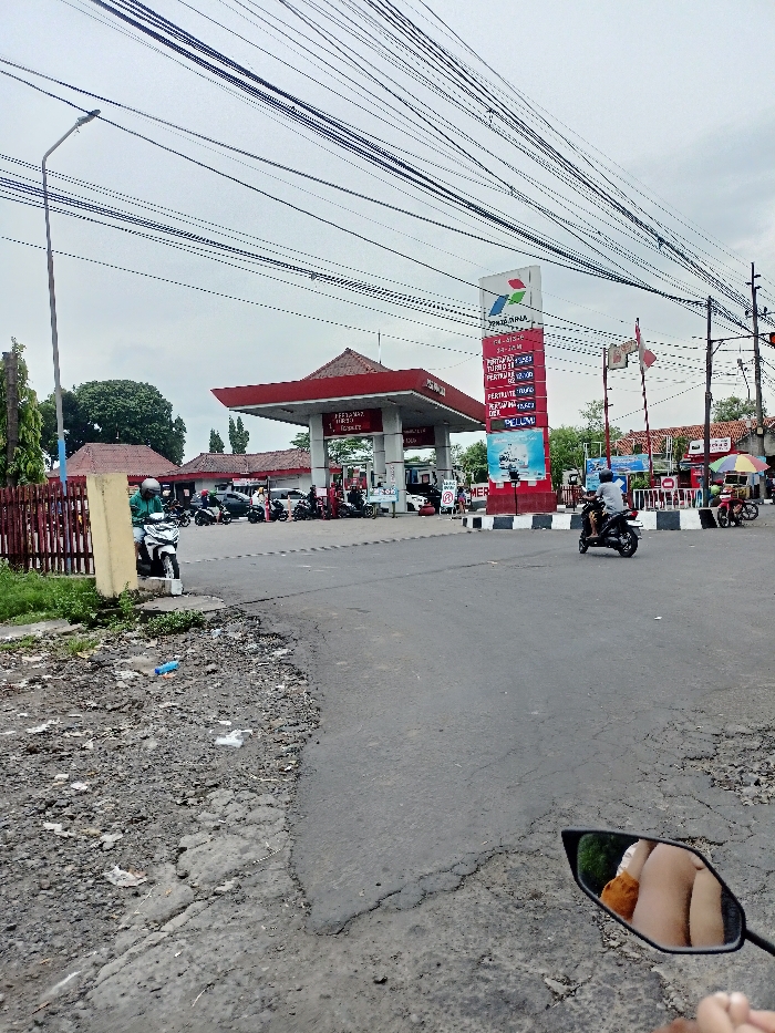 This outdoor medium shot captures a lively street scene in Kecamatan Mojosari, Indonesia, focusing on a Pertamina gas station (SPBU) under an overcast sky. The time of day appears to be daytime, possibly late morning or afternoon, given the lack of strong shadows.
The central feature is the Pertamina gas station, identifiable by its red-roofed canopy and a prominent red and white sign. The sign displays the "PERTAMINA" logo and the phrase "PASTI PAS," along with "HARGA KESEHARIAN" (daily prices). Listed fuel prices include Pertamax (12.500), Pertalite (10.000), Pertamina Dex (13.200), and Dexlite (12.150). Text like "BELILAH DI SPBU" (Buy at SPBU) and an "INDONESIA ENERGI MARITIM" graphic are also visible on the sign.
Multiple people on motorcycles are present at the gas station, either fueling their vehicles or waiting to do so, indicating routine daily activity. On the main road, a motorcyclist in a helmet is seen driving past the gas station. Another person on a white scooter is stationary near a wooden fence on the left side of the frame.
The foreground features a weathered asphalt road transitioning to a rough dirt and gravel shoulder on the left, with some litter present. A tall street light pole stands on the left, and numerous overhead utility wires crisscross the sky. Across the street from the gas station, other buildings and small market stalls with colorful umbrellas can be seen in the background.
A distinct detail is the reflection in the motorcycle mirror in the bottom right corner, revealing the legs and feet of the person taking the photograph, providing a first-person perspective of the scene. The red and white Indonesian flag visible near the gas station entrance further confirms the location. The scene depicts a typical, bustling urban environment in Indonesia.