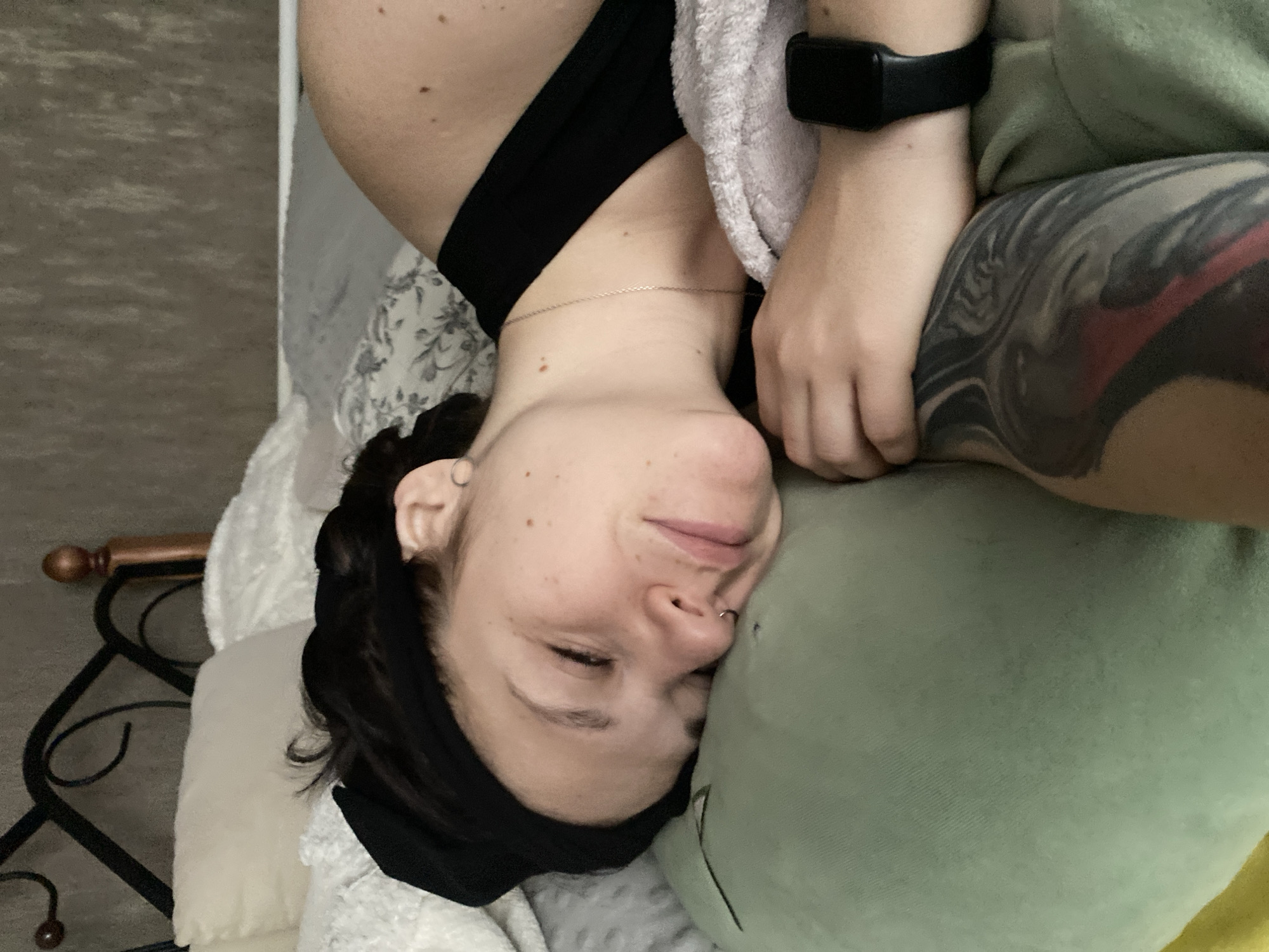 A person with dark hair, fair skin, and a nose piercing is lying in bed, resting their head on a light green pillow. They are wearing a black top and a black fitness tracker on their wrist. A portion of their arm is visible, featuring a large, gray and red tattoo. A white towel is draped near their neck. The bed is covered with various white and gray pillows and blankets, and a black metal bed frame is visible in the background. The location is unspecified, and there is no indication of Chaikovsky, Russia.