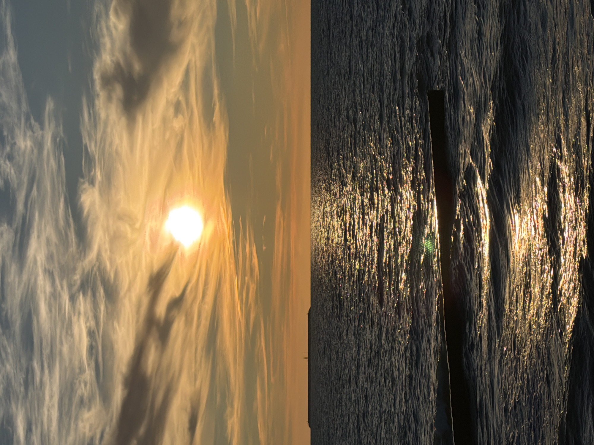 An outdoor, vertical, full shot captures a body of water at sunset. The sun, a brilliant white orb with a faint orange halo, is situated in the upper center of the frame, casting a shimmering golden path across the rippling dark water. Wispy, light-colored clouds streak across the sky, tinged with orange and yellow from the setting sun. On the horizon, a long, flat structure, possibly a pier or breakwater, extends across the water from left to right, with a small lighthouse visible on its far left end. In the foreground, a dark, partially submerged object, possibly a piece of driftwood or debris, lies horizontally on the surface of the water. The scene is quiet and serene, bathed in the warm glow of the sunset. This view is typical of the Baltic Sea coast in Zelenogradsk, Russia, a popular resort town known for its beaches and maritime landscape.