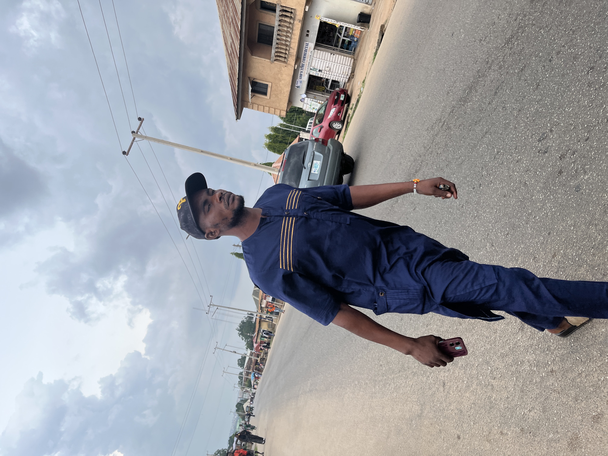In Jos, Nigeria, a young man walks down a street holding a smartphone. He wears a dark blue, embroidered tunic and matching trousers, and a black baseball cap. The man is positioned in the foreground, capturing his side profile and the lower half of his body. Behind him, the street stretches into the distance, lined with shops and buildings. A grey SUV is parked on the right side of the road, and a red car is visible further down. The sky is overcast with dramatic clouds.