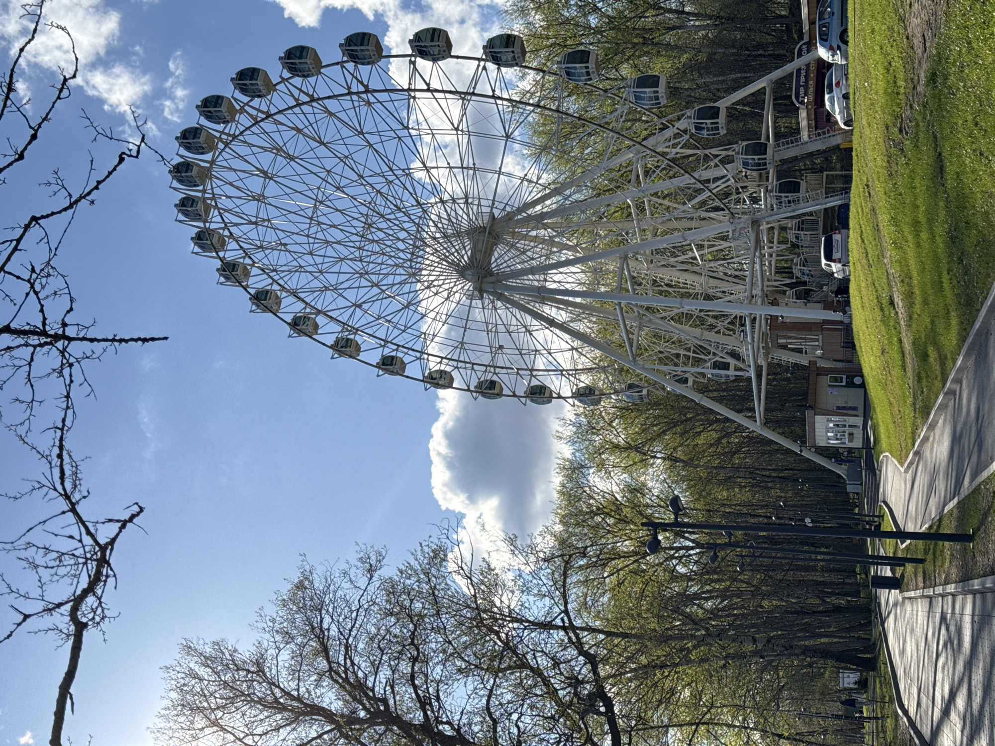 A tilted low-angle shot of a Ferris wheel against a blue sky with scattered clouds. The Ferris wheel is white and features numerous passenger cars evenly spaced around its circumference. Trees surround the base of the wheel, and some bare tree branches frame the left side of the image. To the right, a paved pathway runs alongside a grassy area. Several parked cars and small buildings are visible in the background. The image shows the Ferris wheel in Park Pobedy in Cherepovets, Russia.