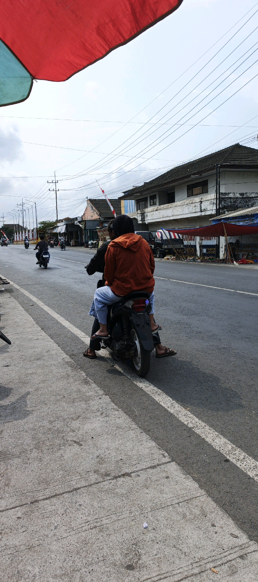 Two individuals are seated on a black motorcycle, viewed from behind, traveling on a paved road. The person in the rear is wearing an orange hooded jacket and light blue pants, while the person in front is wearing a dark top. Both are wearing sandals. The motorcycle has a visible license plate mount. They are on a road with two lanes of traffic, marked by a white line. To the left of the road is a concrete sidewalk or pedestrian area. In the background, there are buildings with tiled roofs, some of which appear to be shops or houses. Overhead, power lines are strung between poles. A red and white striped barrier is visible in the distance, possibly indicating a checkpoint or a festive decoration. A large red and teal colored fabric, likely an umbrella or canopy, is partially visible in the upper left corner of the frame. The sky is overcast. The scene appears to be in Kasiyan, Indonesia, given the context.