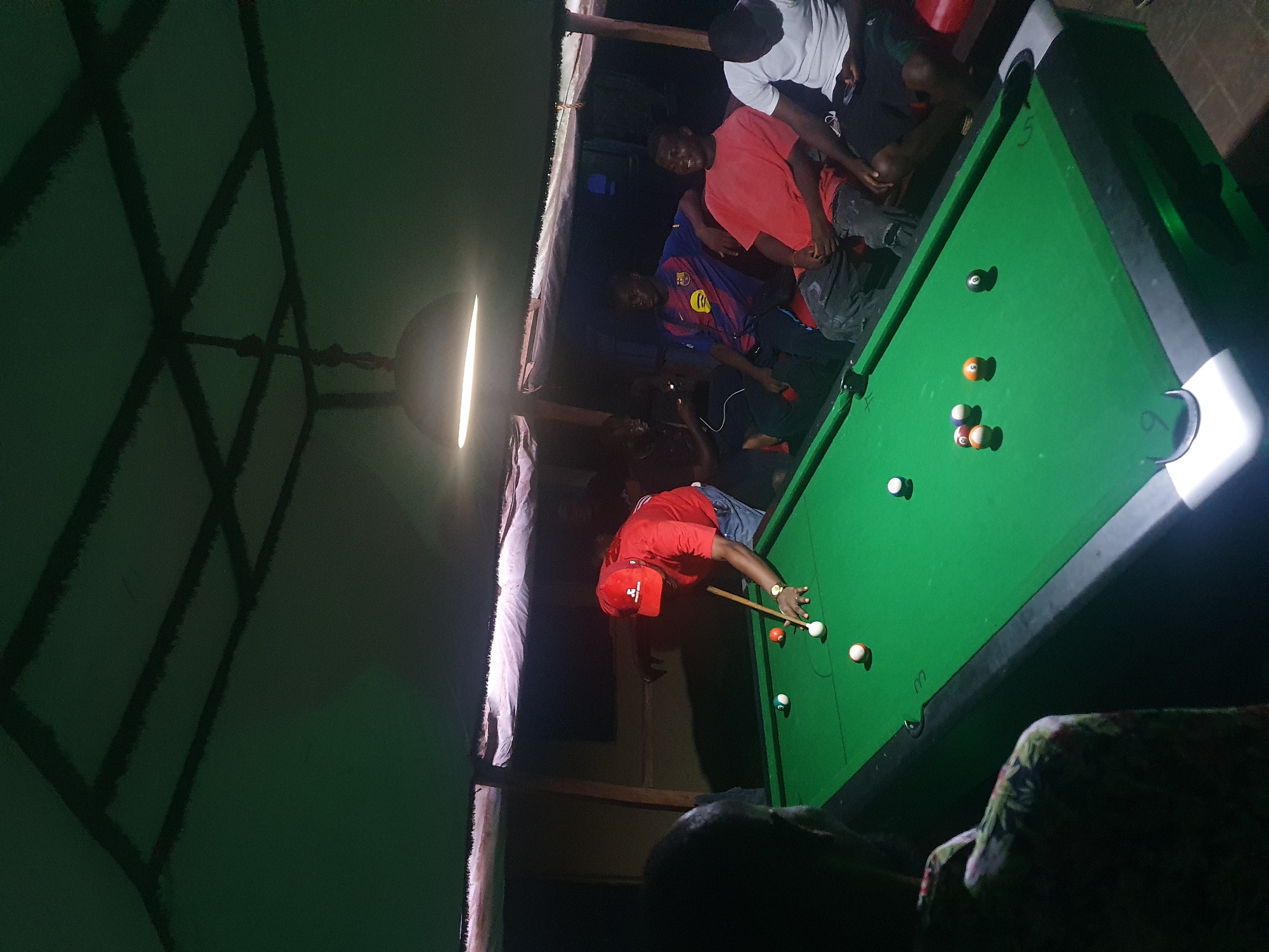 The image depicts an indoor scene at night in Lokoja, Nigeria, showing a group of men gathered around a pool table. The central figure, wearing a red t-shirt, blue jeans, and a red baseball cap, is actively playing pool, leaning over the green felt table with a cue in hand to take a shot. A gold-colored watch is visible on his left wrist.
Several pool balls are on the table, including the white cue ball, a solid black 8-ball, a solid orange ball, and other striped and solid balls. A bright, round overhead light fixture illuminates the pool table, casting strong shadows and highlighting the activity.
Other men are observing the game. One man in a blue and red striped FC Barcelona jersey stands near the table, while another in a white t-shirt and black shorts is seated or leaning on the opposite side. Additional indistinct figures are visible in the dimly lit background. The setting appears to be a casual local establishment, possibly a pool hall or bar, characterized by dark wooden structural beams in the ceiling. The overall lighting is dim, emphasizing the brightly lit pool table. Faint numbers "5" and "6" are visible on the edge of the pool table near two pockets. A piece of patterned furniture is partially visible on the right side of the frame. The atmosphere is social and relaxed.