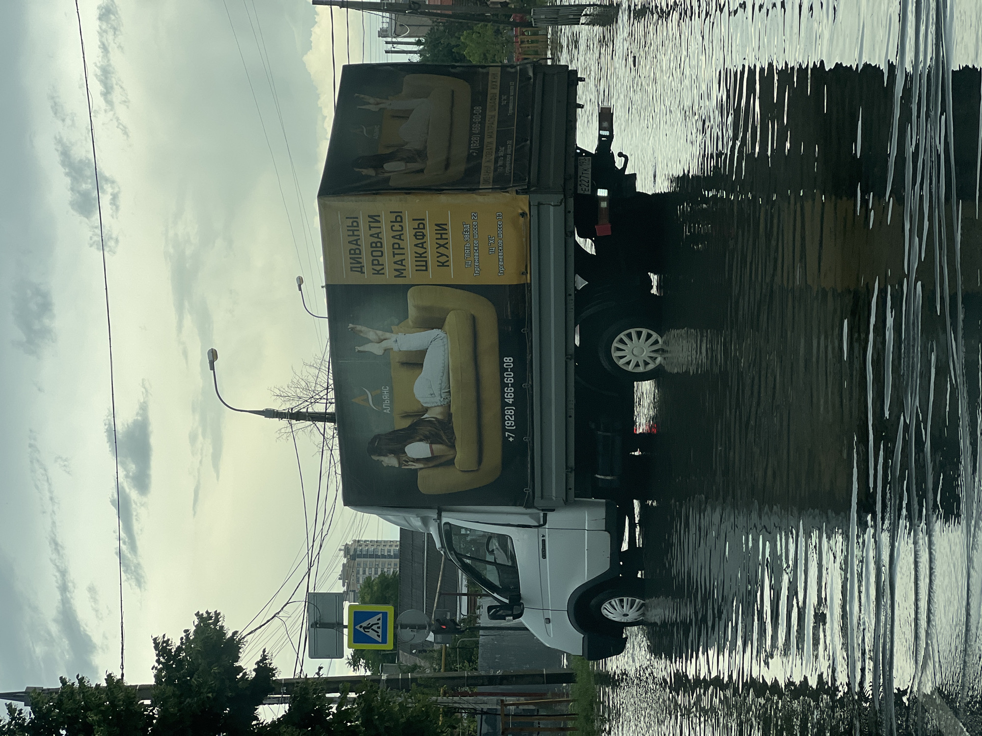 A white truck is driving through a flooded street in Krasnodar, Russia. The truck is a delivery truck with an advertisement on its side for furniture, including sofas, beds, mattresses, wardrobes, and kitchens. The advertisement features images of people lounging on sofas. The street is covered in several inches of water, indicating heavy rainfall or a plumbing issue. The sky is overcast and grey, suggesting that the weather is poor. Other objects visible in the image include a traffic light, a street sign with a pedestrian crossing symbol, trees, and power lines. The truck's license plate is partially visible and reads "E227TK 193". The reflection of the truck and the surrounding environment can be seen on the surface of the flooded street.