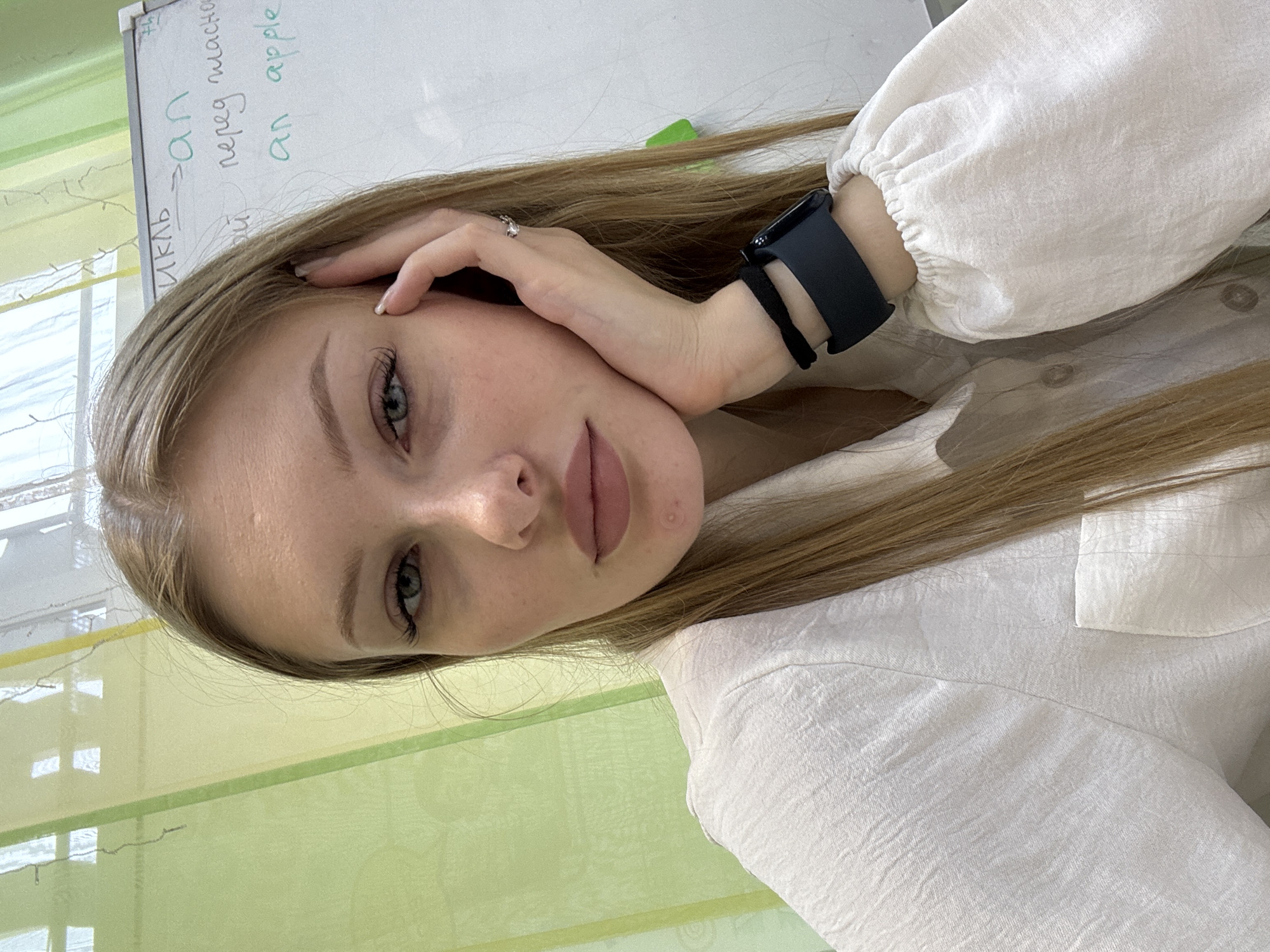A young woman with long, blonde hair is looking at the camera. She is wearing a white, long-sleeved blouse with a textured fabric and a round neckline. A dark smartwatch is visible on her left wrist. Her left hand is resting on her cheek, and she is wearing a silver ring on her index finger. Her makeup includes mascara, eyeshadow, and lip gloss. A pimple is visible on her chin.
Behind her, a whiteboard is partially visible, with handwritten text in Russian and English, including "цикл", "an", and "an apple". The background appears to be an indoor setting with green and yellow vertical blinds or curtains. The overall lighting suggests an indoor environment, possibly a classroom or office.
The image is a selfie taken from a slightly low angle. While the image suggests an indoor educational setting, there are no specific visual cues to pinpoint the exact location within the city of Perm, Russia.