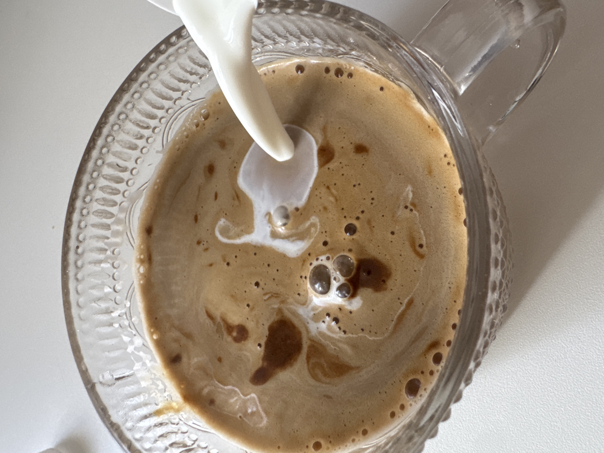A close-up, overhead shot shows milk being poured into a clear glass mug filled with coffee. The coffee has a frothy, tan-colored surface with darker brown swirls and small bubbles. The milk is a creamy white liquid and is creating ripples and patterns as it mixes with the coffee. The mug has an ornate, textured rim. The background is a plain, light-colored surface, likely a table. The image does not contain any information that would allow for the identification of a specific location in Chaikovsky, Russia.