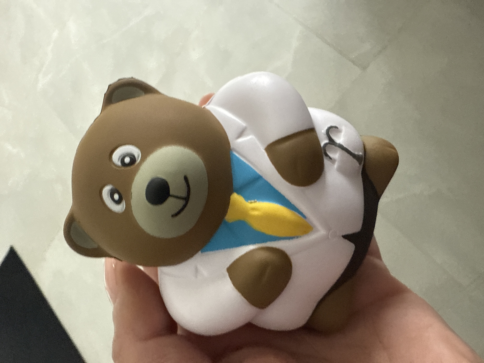The image depicts a hand holding a toy bear dressed as a doctor. The bear is brown with a white coat, blue shirt, and yellow tie. The background appears to be a tiled floor and a light-colored wall. The location cannot be determined to be in the city of Davie, United States of America without further information.