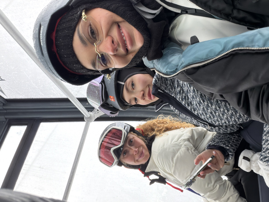 Three women are seated inside a ski lift gondola cabin, likely in Les Allues, France, during snowy or overcast weather. The woman on the left has curly blonde hair, wears a white puffer jacket, black ski helmet with red goggles pushed up, and holds a silver smartphone in her right hand. She smiles towards the camera. The woman in the center wears a black and white patterned jacket and a black ski helmet with iridescent goggles pushed up; she looks towards the camera with a slight smile. The woman on the right, partially visible, wears a black and blue ski jacket, a black ski helmet with an "ATOMIC" logo, and gold-rimmed glasses with tinted lenses. She is smiling directly at the camera. All three are dressed in winter ski apparel. The exterior view through the cabin window is obscured by precipitation and an overcast sky.