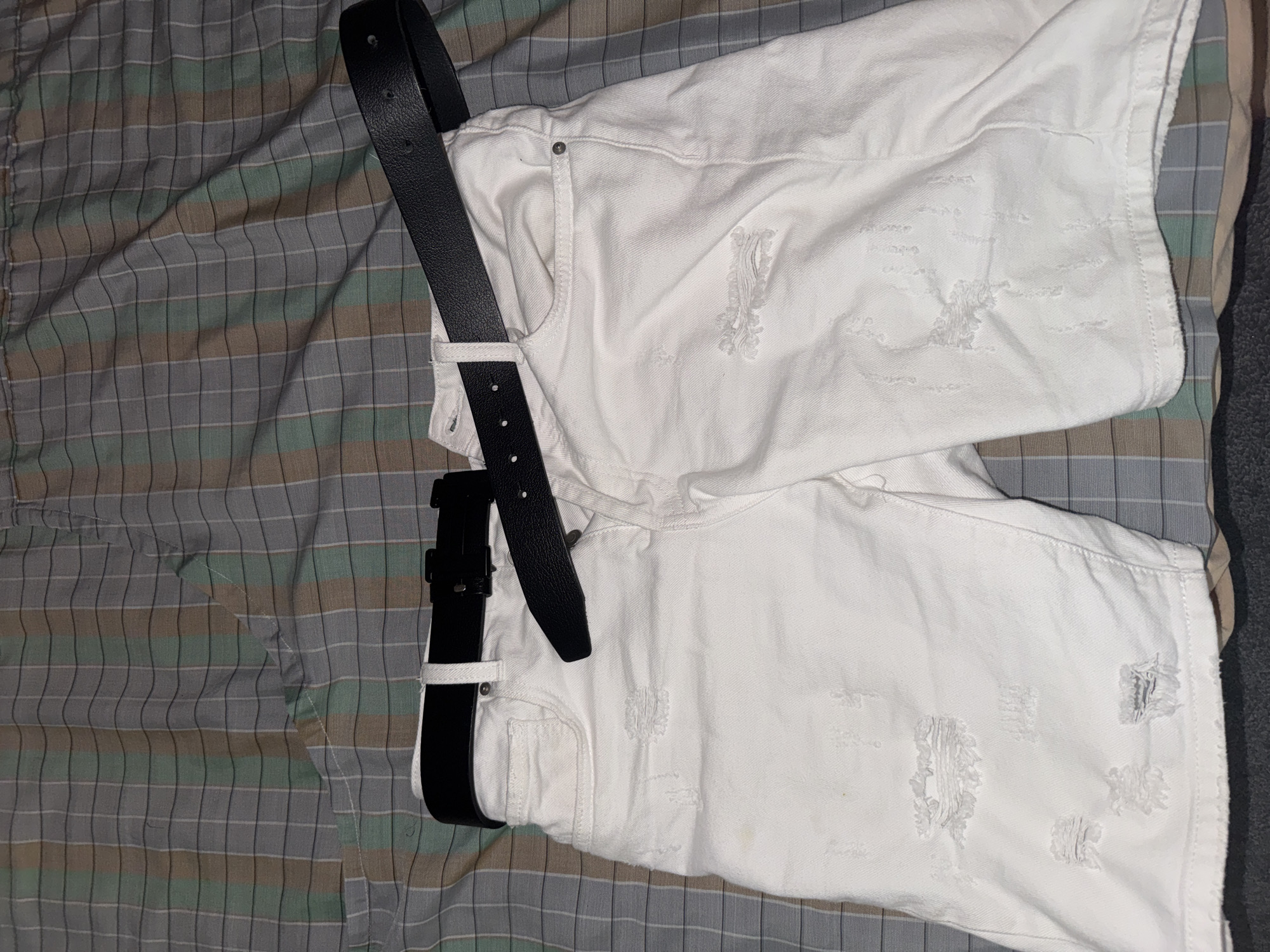The image shows a pair of white denim shorts with distressed detailing, placed on a patterned fabric. A black leather belt with a silver buckle is draped across the shorts. The image does not contain any information that would allow it to be located in Ruse, Bulgaria or any other location. There are no people depicted in the image.