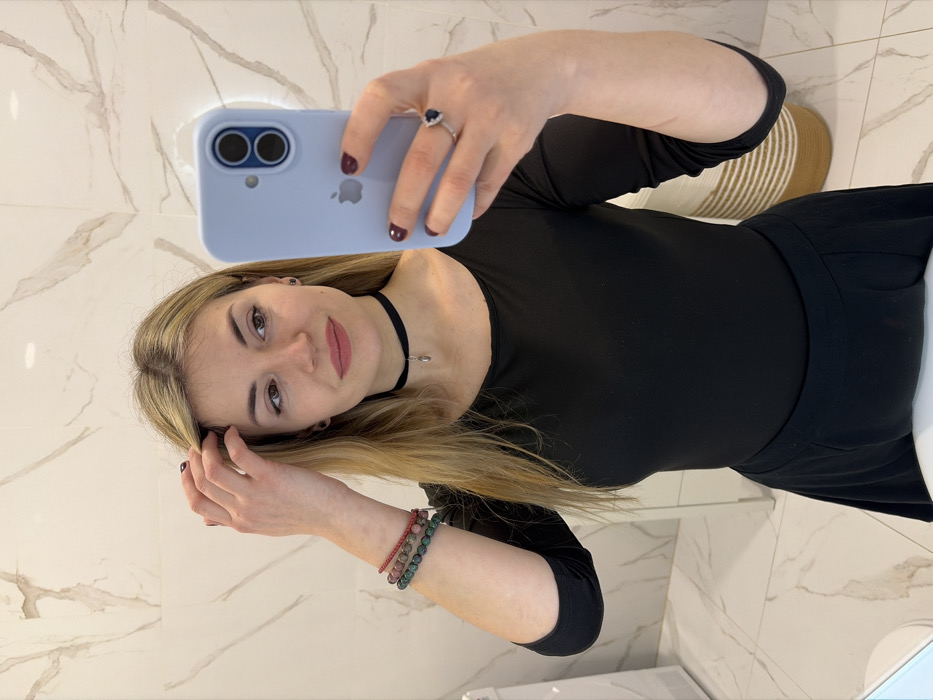 A female individual is depicted from the waist up, taking a mirror selfie indoors. She has long, blonde hair, light-toned skin, and is wearing a black, scoop-neck, three-quarter sleeve top and a black skirt. Her left hand is raised, with fingers touching her hair on the left side of her head. Adorning her left wrist are three beaded bracelets: one red, one multi-colored green and red, and one green. On her left ring finger, she wears a silver-colored ring featuring a prominent dark blue oval-shaped stone surrounded by smaller clear stones. A black choker with a silver teardrop-shaped pendant is worn around her neck. Her fingernails are painted a dark red. She holds a light blue Apple iPhone, visible by its logo and dual camera module, in her right hand, pointed towards the mirror. The background consists of light-colored tiles with a subtle grey marble-like veining pattern on both the wall and floor. Partially visible objects include a white fixture to the far left and a woven storage basket to the right. The scene is located within an indoor setting in the city of Budva, Montenegro.
