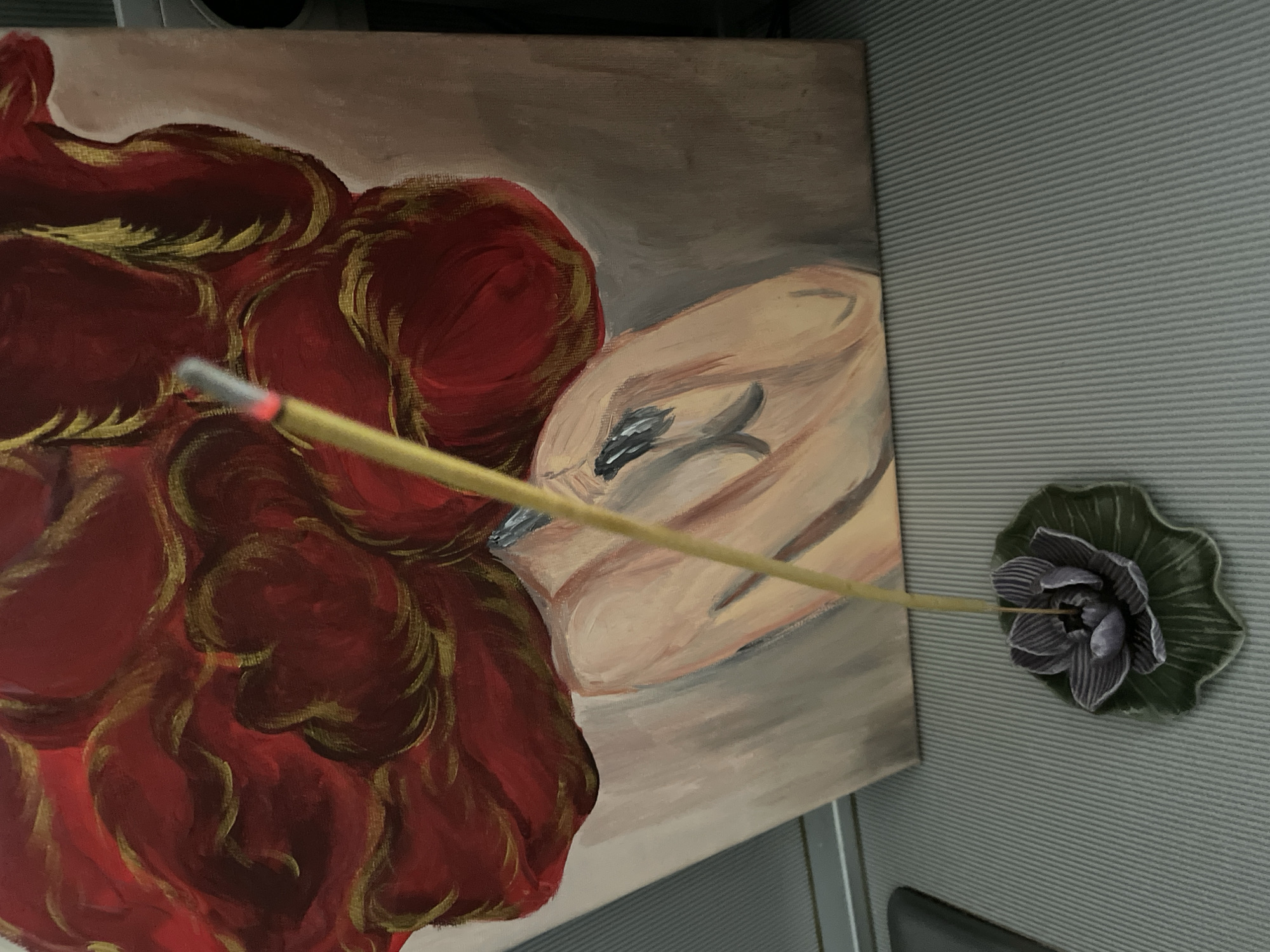 The image shows an indoor scene with a painting and an incense stick. The painting depicts a person with a large, voluminous mass of red and gold hair, with parts of their shoulders and neck visible. The person's skin tone is rendered in shades of peach and pink. The background of the painting is a muted, light brown. In the foreground, an unlit incense stick stands upright in a dark purple lotus-shaped incense holder, which is resting on a green leaf-shaped base. The incense stick is positioned to the left of the painting. The scene is set on a gray, ribbed surface. The location of this scene in the city of Sochi, Russia, cannot be determined from the provided image.