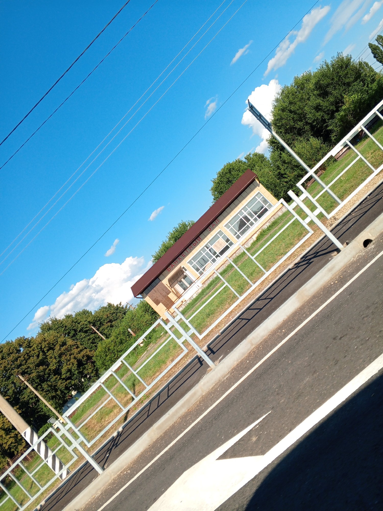 The image shows a view of a building with a brown roof and yellow facade, featuring large windows. The building is situated next to a grassy area with a white metal fence in the foreground. A road is visible in the lower right of the frame, with white lane markings. Overhead, power lines are strung against a bright blue sky with scattered white clouds. Lush green trees line the background. The image is taken at an angle, giving it a dynamic perspective. The sign on the building reads "БАНКЕТНЫЙ ЗАЛ," indicating it is a banquet hall. This scene is likely located in Pervomaiskoye, Russia, given the context of the provided information.