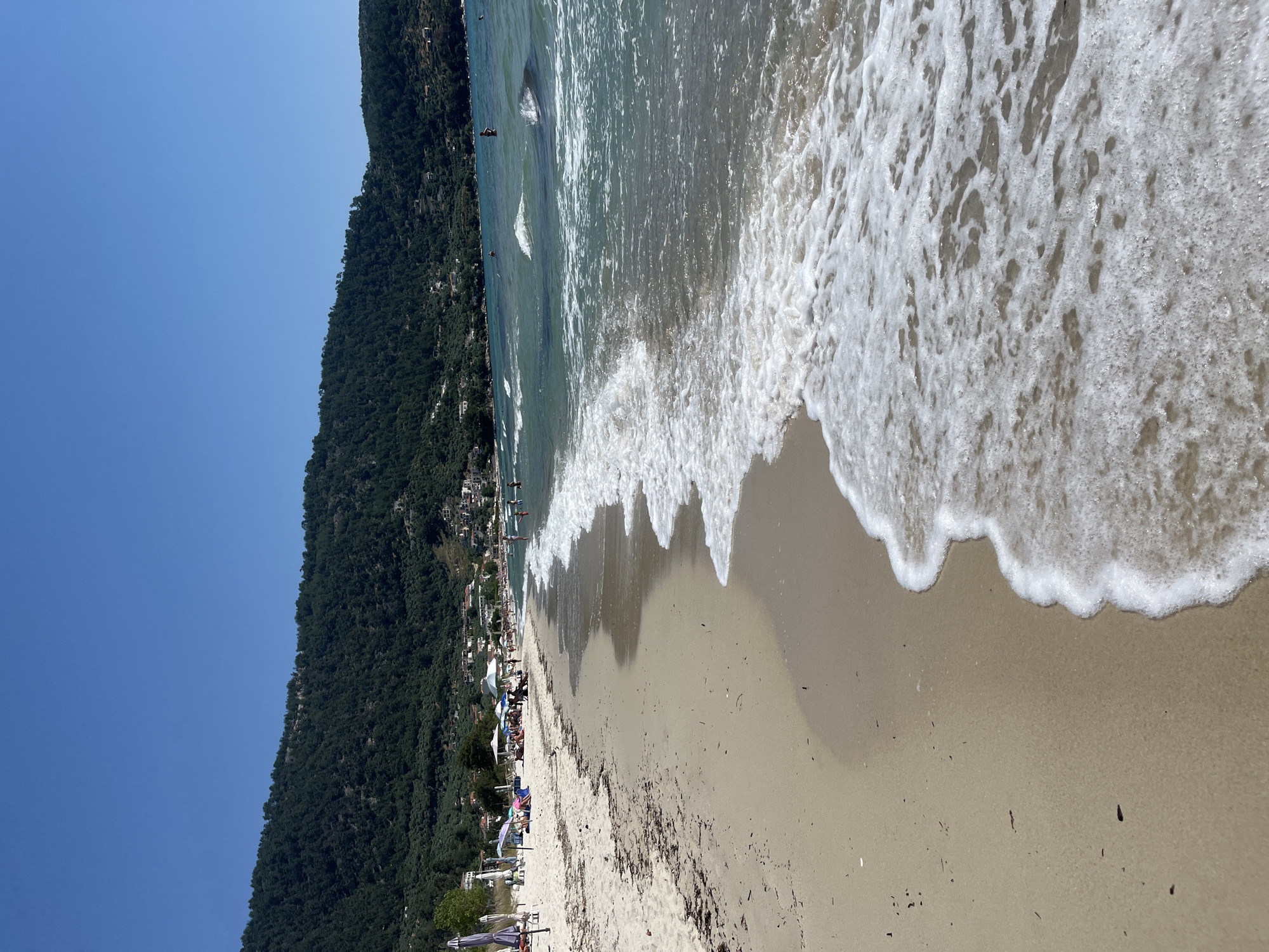 This is a photograph of a sandy beach with clear blue water and a lush green hillside in the background. The sky is a solid, bright blue. On the beach, there are several umbrellas and beach chairs, with a few people sunbathing and walking. The water is calm with small waves gently lapping the shore. In the distance, a town is visible nestled on the hillside. The overall scene depicts a peaceful and sunny day at a coastal resort.