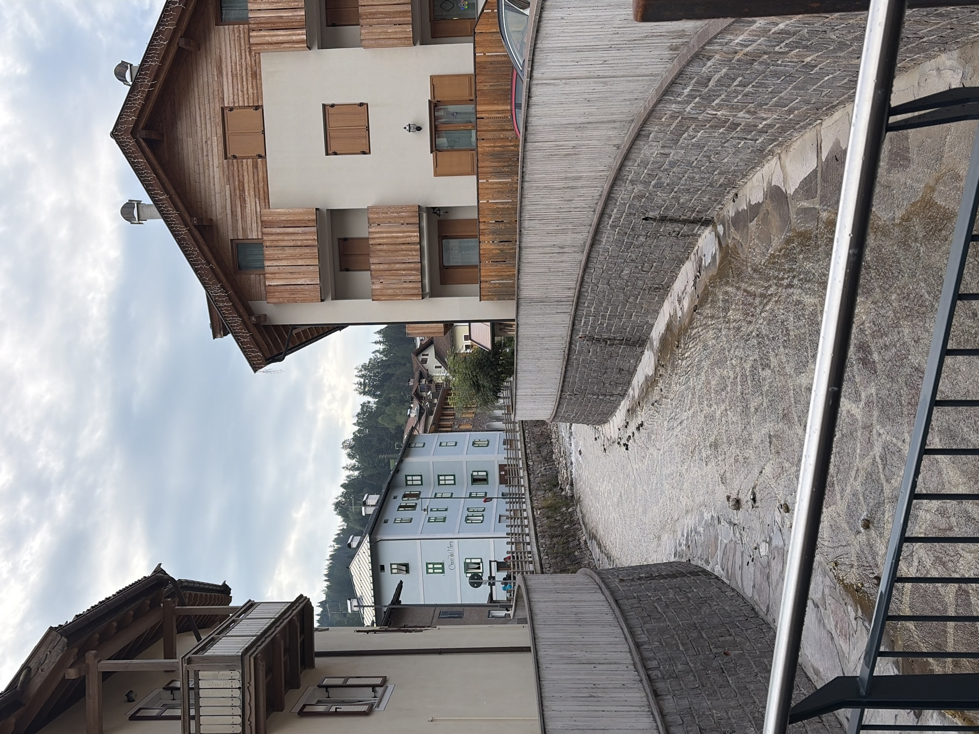 The image shows a street scene in Moena, Italy, with a canal running through the center of the frame. The canal is bordered by stone walls and has a shallow, fast-moving stream of water with visible rocks and debris. Buildings line the sides of the street, with a focus on traditional alpine architecture. On the left, a light-colored building features a wooden balcony with white railings. To the right, a taller building with a wooden facade and multiple balconies with wooden railings is visible. Further in the background, a light blue building with green trim is identifiable, displaying the text "Casa del Moro". The sky is overcast with clouds. There are no discernible people actively engaged in interaction within the immediate frame. The scene depicts an urban waterway bordered by architecture in a town setting.