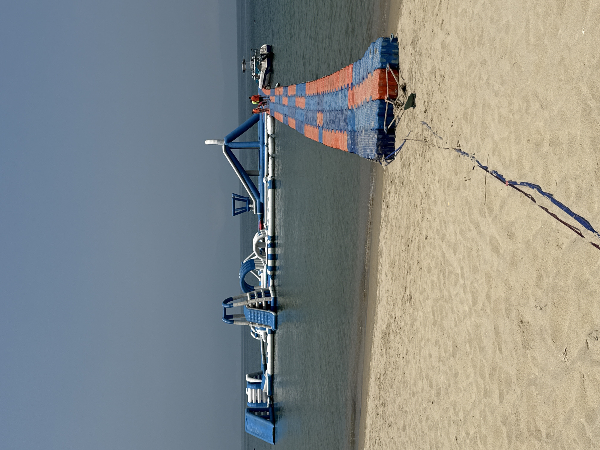 The image depicts a beach scene in Đà Nẵng, Vietnam, featuring a large inflatable water park situated in the calm ocean water under a hazy sky. In the foreground, a sandy beach extends to the waterline. A floating walkway, constructed from alternating blue and orange plastic modules, originates from the beach and extends into the water towards the inflatable park. Three individuals, two in orange tops and one in a yellow/green top, are walking on this walkway towards the water park. A small, white powerboat with a blue stripe is moored to the right of the walkway, near the inflatable structure. The inflatable water park itself is predominantly blue and white, comprising various slides, climbing elements, and interconnected platforms. Distant mountains are faintly visible on the horizon beneath the light blue-grey sky. Ropes secure the floating walkway to the sand in the foreground.