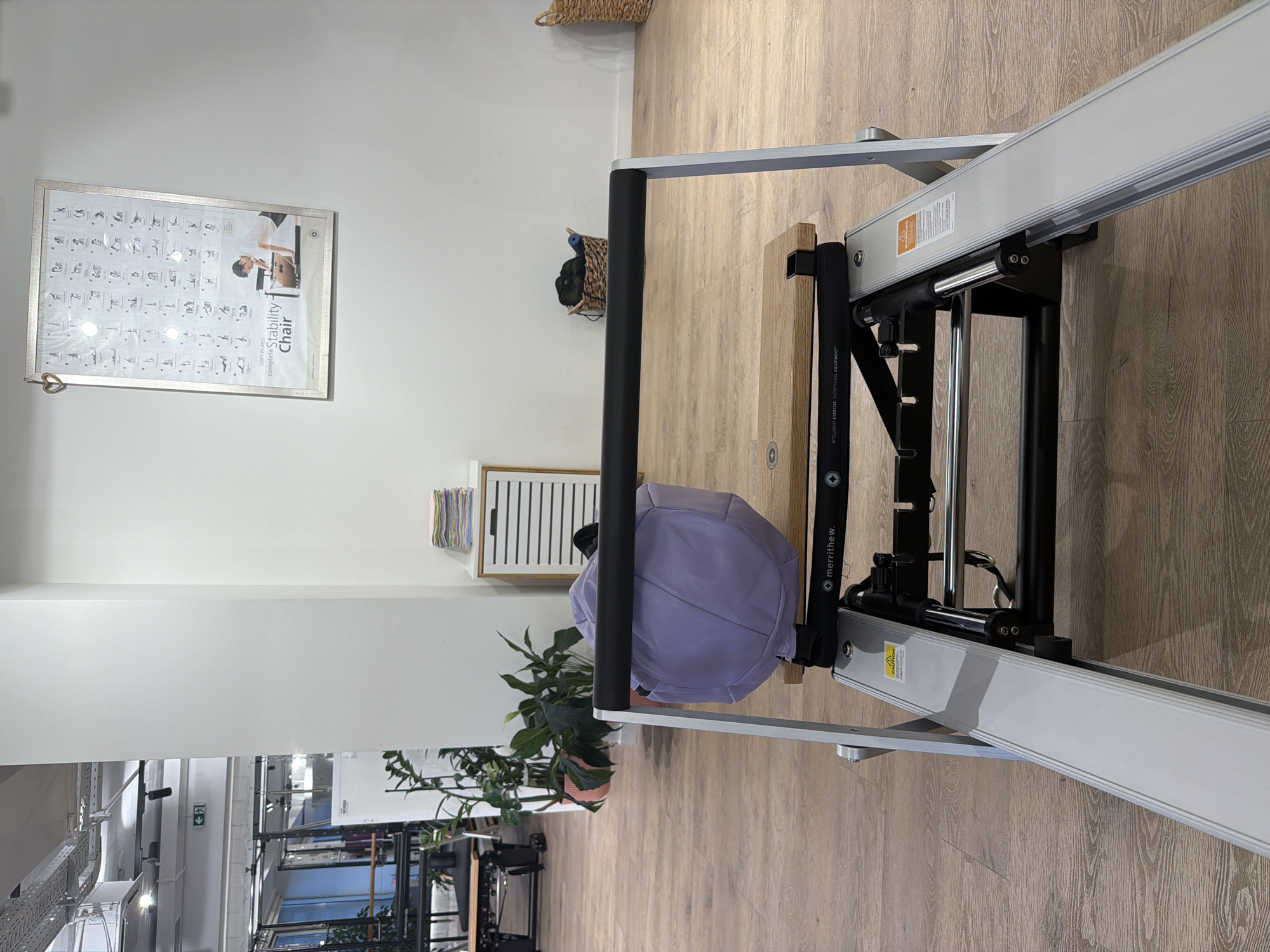 The image depicts the interior of a fitness studio, likely a Pilates studio, as indicated by the equipment and poster. The scene is viewed from a low angle, looking across a Pilates reformer machine. On the reformer, there is a light purple duffel bag. Behind the reformer, on the wooden floor, are two baskets, one containing dark green items and the other is empty. To the right, a woven basket is visible. Further back, a white cabinet with horizontal slats is present, topped with a stack of colorful folded towels. A large framed poster hangs on the white wall, illustrating exercises for a "complete Stability Chair" by STOTT PILATES. Plants in pots are visible to the left, along with other gym equipment and exposed ceiling infrastructure, suggesting a functional and active space. The floor is a light-colored wood laminate. The location is specified as Boreti, Montenegro. There are no people visible in the image.
