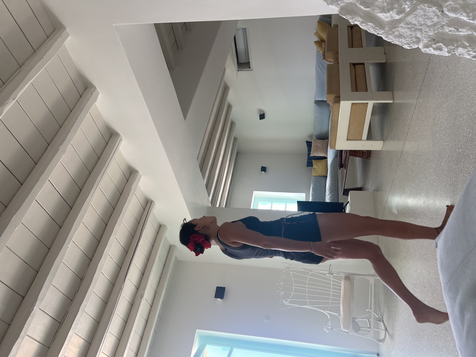 The image displays the interior of a brightly lit residence located in Tourlos, Greece, characterized by white-painted walls, a light-colored floor, and a white wooden beamed ceiling. A single female individual, appearing to be of Asian descent, is observed in profile walking barefoot from left to right across the foreground. She is dressed in a black tank top and black shorts, and her dark hair is adorned with prominent red and black floral accessories.
To the left, a white, ornate metal rocking chair with a light-colored cushion is positioned. The background reveals a living area featuring a large, light blue L-shaped sectional sofa accessorized with various blue, yellow, and one pink pillow. A dark wooden coffee table is centered in front of the sofa, flanked by two light-colored armchairs with yellow cushions. A television is visible on a low white stand to the left of the sofa. The back wall includes two dark wall-mounted speakers, a small square window with a light blue frame, and a mounted air conditioning unit. Another larger square window with a light blue frame is present on the far left wall. An open archway is visible in the background, leading to an adjacent area within the dwelling.