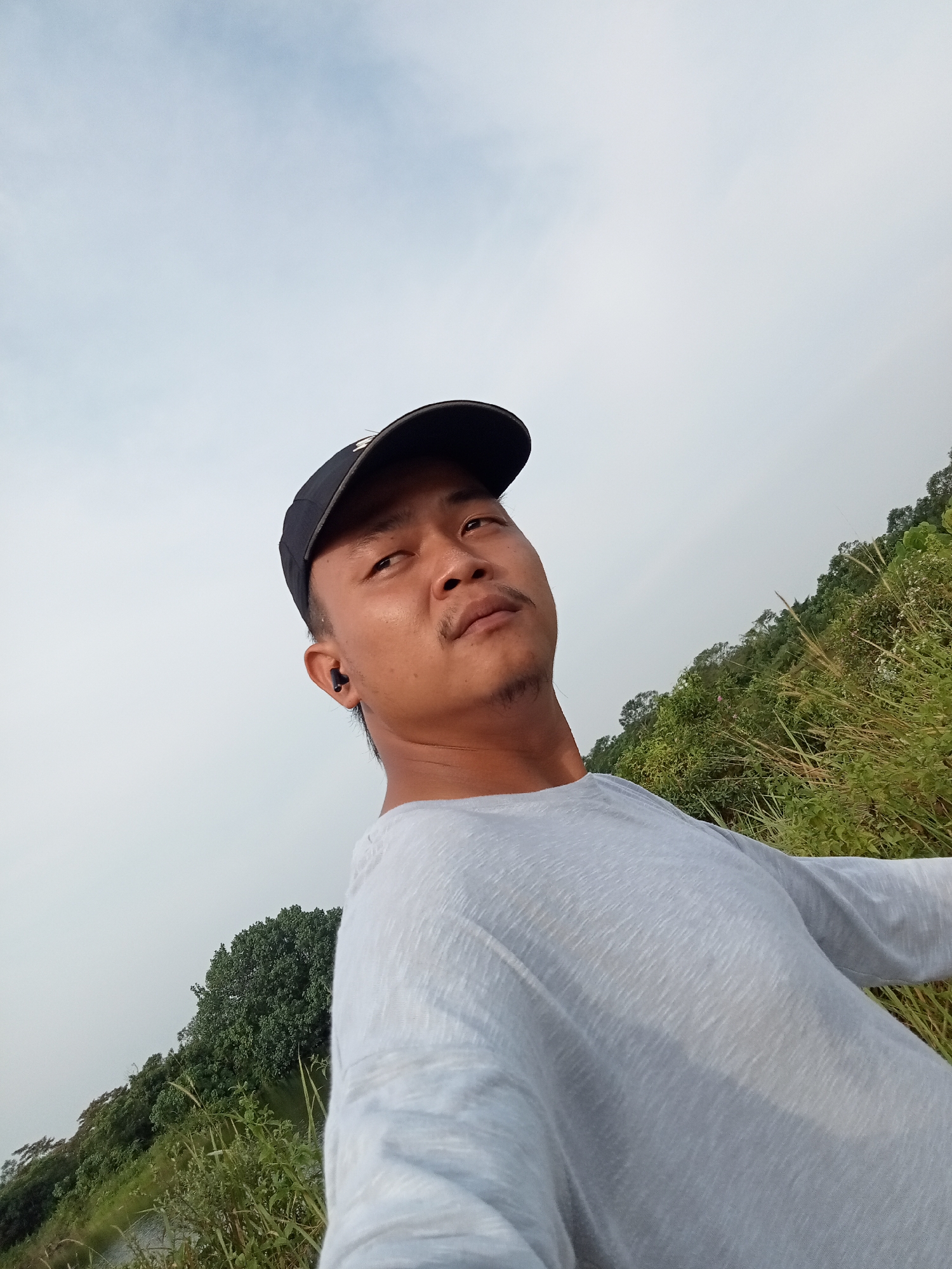 A man wearing a light gray long-sleeved shirt and a black baseball cap is captured in a selfie. He has an earbud in his left ear. The background consists of a lush green hillside covered in trees and tall grass, under a pale sky. The scene appears to be an outdoor environment, possibly a rural area near Kualu, Indonesia, with the natural landscape suggesting a location away from the immediate urban center.