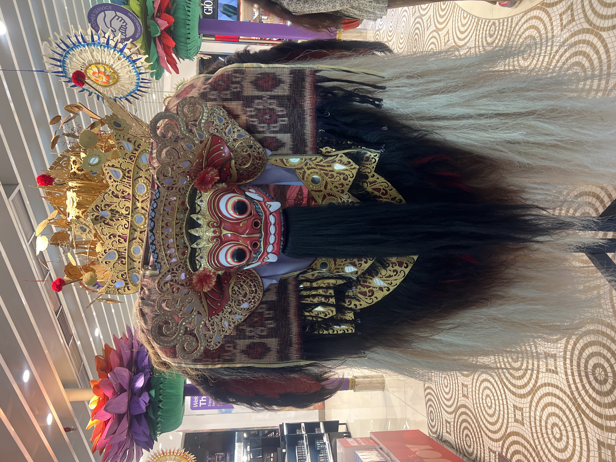 A Barong mask, an iconic symbol of Balinese culture, is on display indoors. The mask features an ornate golden crown, a red face with large eyes, and a long black beard. A patterned cloth covers the head, and long strands of black and white hair extend from the back. Artificial lotus flowers hang nearby. The mask is located in a shopping center in Kuta, Indonesia.