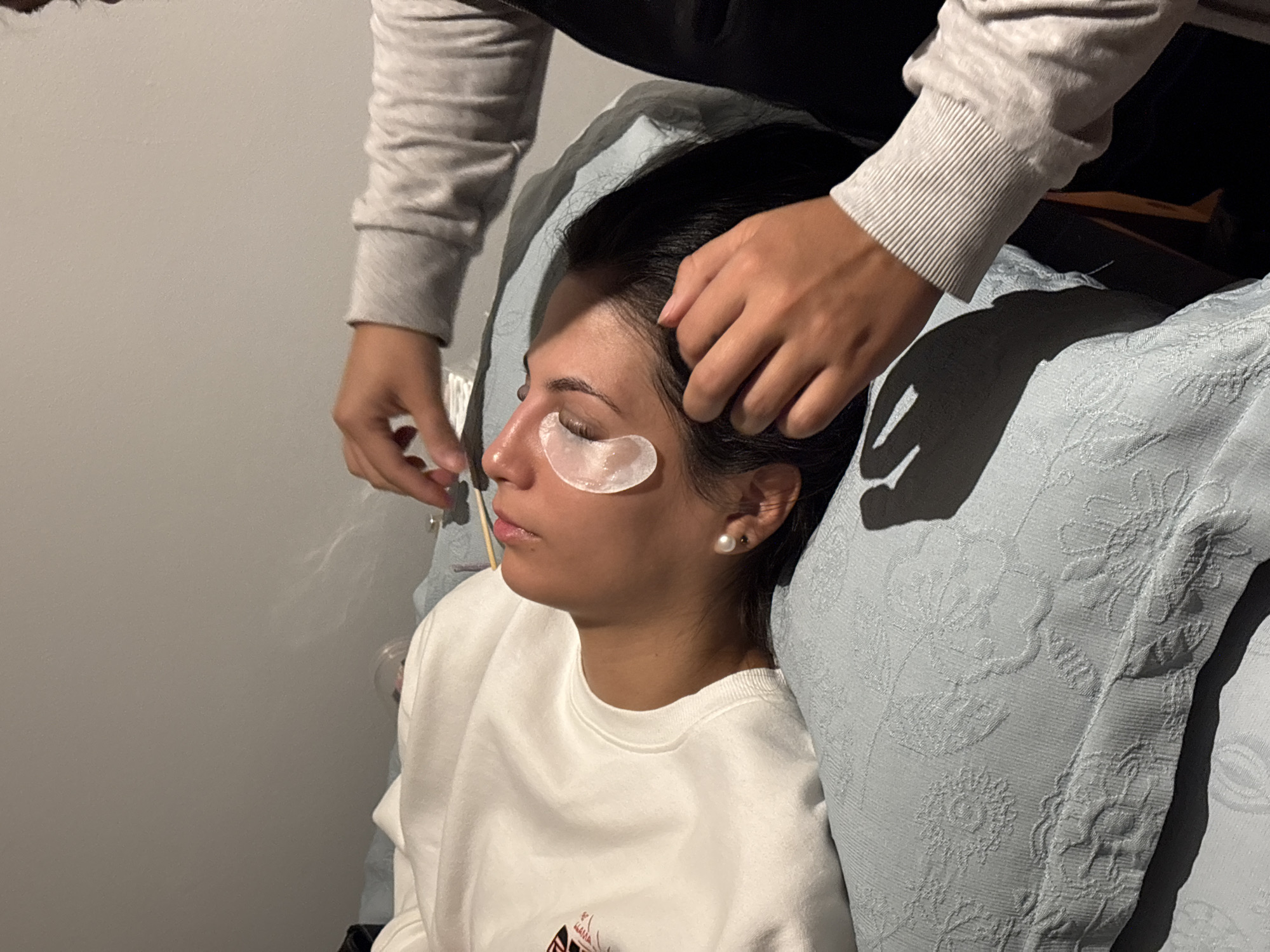A person is lying down, facing upwards, with their eyes closed. Underneath their eyes are two white under-eye patches. A second person, positioned above and to the right, is holding a small applicator with a pointed tip and appears to be working near the first person's eyes, possibly applying eyelash extensions. The scene takes place indoors, on a bed covered with a light blue patterned blanket. The walls are a neutral gray. The lighting is dim. The location is not specified in San Isidro, Peru.
