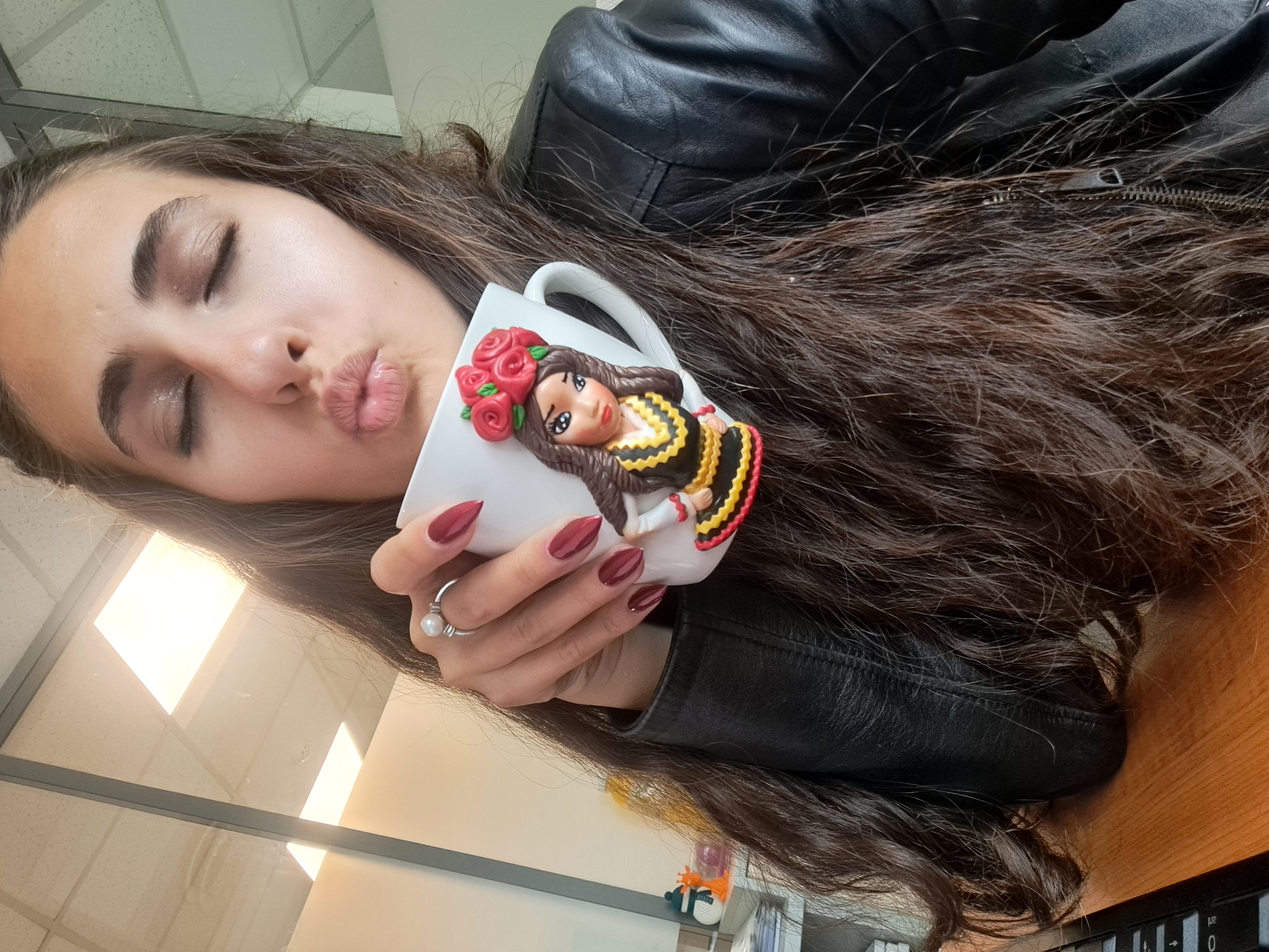 A woman with long brown hair and dark clothing is posing for a selfie with a white mug. She is puckering her lips, her eyes are closed, and she is holding the mug up to her face. The mug has a decoration of a woman with brown hair, a red rose headpiece, and a yellow and black dress. The woman's nails are painted dark red. The background appears to be an office setting with light-colored walls and a desk. There is no explicit information to confirm that this image was taken in the city of Sofia, Bulgaria.