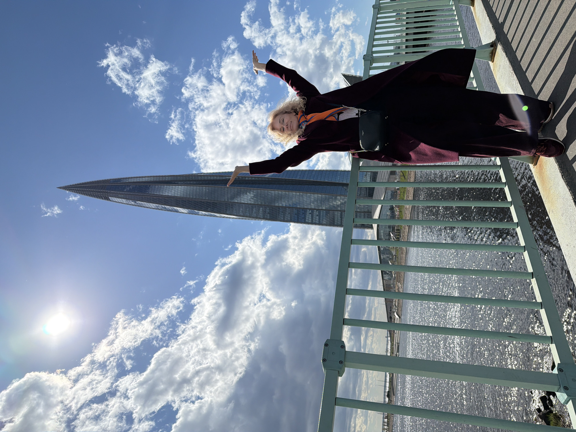 A woman is positioned against the backdrop of a partly cloudy sky and the Lakhta Center skyscraper in Saint Petersburg, Russia. She stands on a bridge with light green railings, extending both arms outward. She wears a long, dark coat, a patterned scarf, and carries a small black purse. The sun shines brightly in the sky. Below the bridge, water reflects the sunlight.