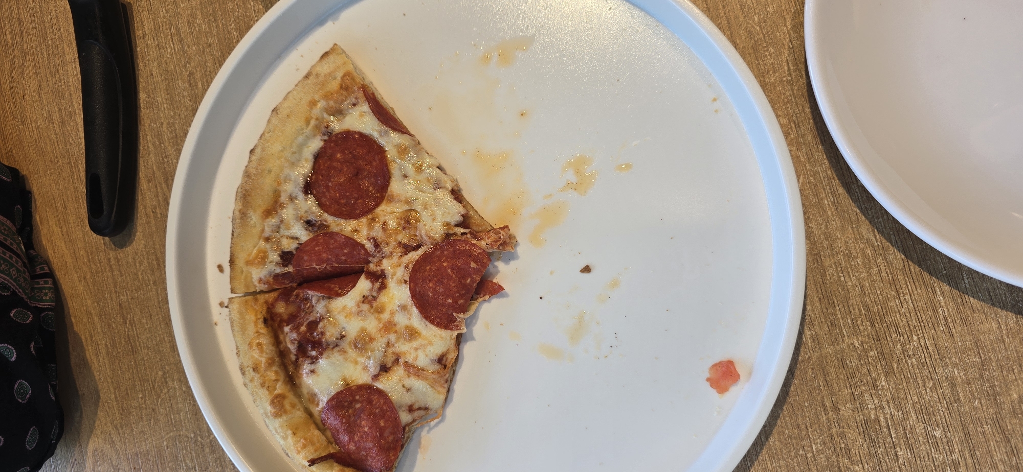 An overhead shot shows two white plates on a wooden table. The plate in the foreground is mostly empty, with a few crumbs and sauce stains. A single slice of pepperoni pizza rests on the right side of the plate. The plate in the background is empty and partially visible at the top of the frame. A black utensil handle is visible in the lower left corner of the image.
