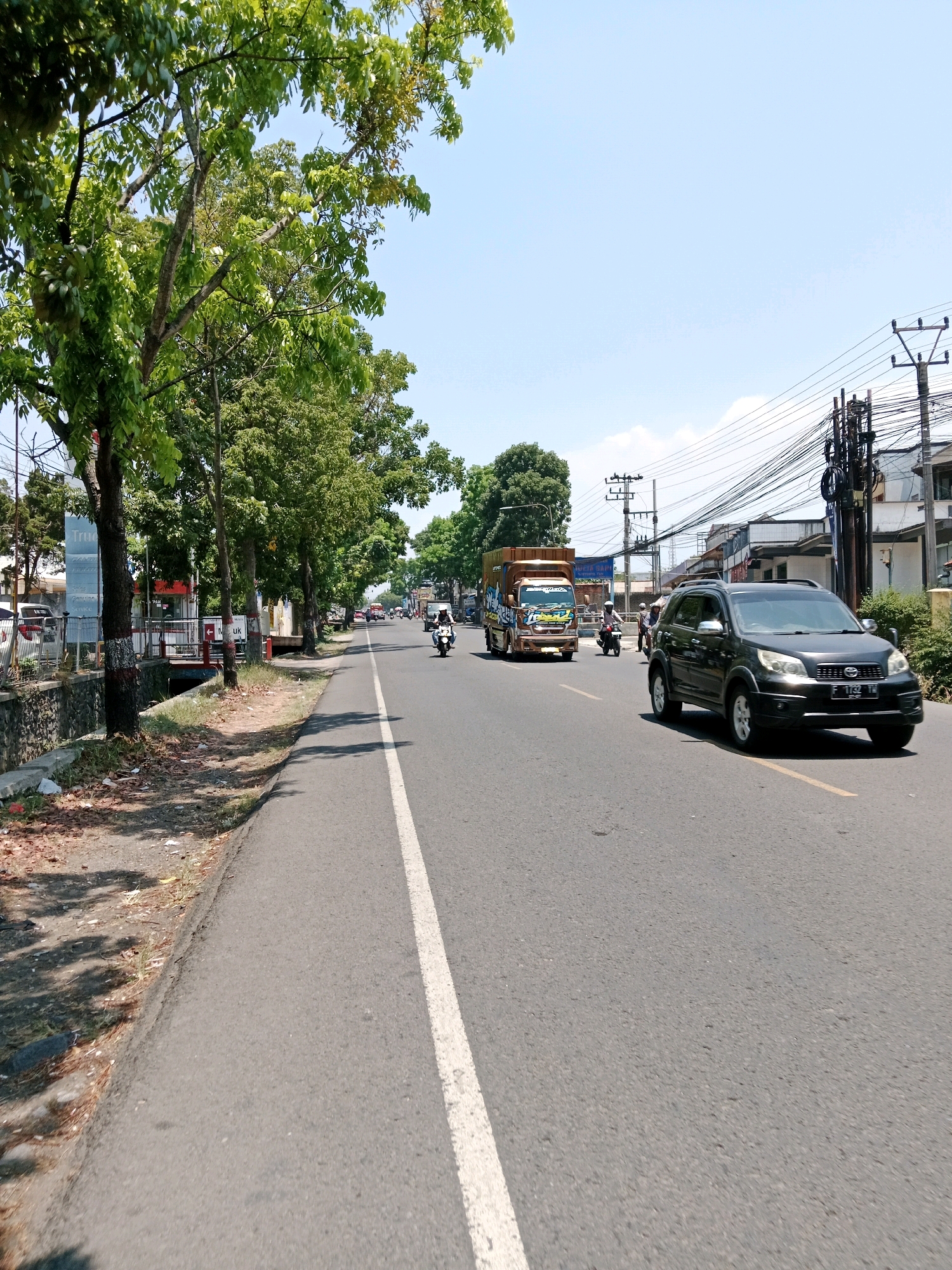 The image depicts a sunny day on a multi-lane road in Cianjur, Indonesia. The road is asphalt with a white dividing line and a yellow line on the right side. Trees line the left side of the road, casting shadows on the sidewalk. Several vehicles are present, including a dark SUV in the foreground, a brown truck with graphics, a motorcycle with a rider, and other vehicles further down the road. Buildings and utility poles with numerous wires are visible on the right side of the road. The sky is clear and blue.