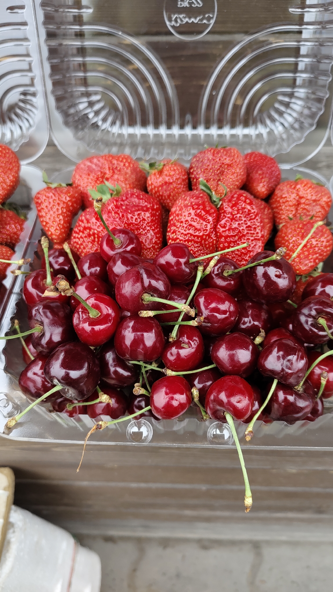 The image shows a clear plastic container filled with a mix of fresh strawberries and dark red cherries. The strawberries are predominantly red with visible seeds and green stems, while the cherries have long green stems. The container appears to be sitting on a wooden surface. The scene is likely at a market or store, but there is no definitive evidence that the location is in the city of Krasnoyarsk, Russia.