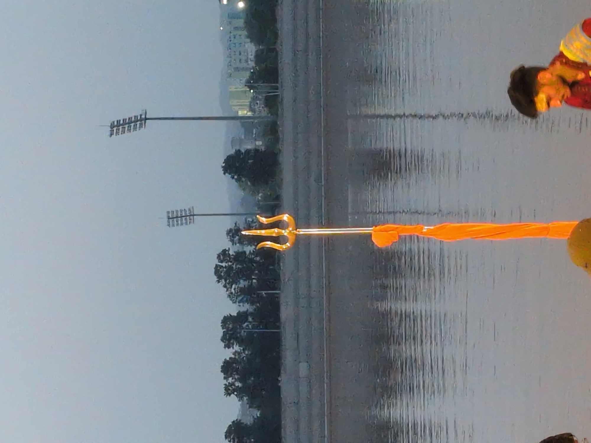 The image captures a scene at the Tawi River in Jammu, India, during dusk or dawn. In the foreground, a golden trident, a religious symbol, stands tall, draped with an orange cloth. The river's surface reflects the muted sky and the surrounding environment. In the background, a concrete embankment lines the river, beyond which are trees and distant buildings. Two tall stadium lights are visible against the sky. On the right side of the frame, a young man with dark hair and a beard is partially visible, wearing a red shirt and a patterned vest. He appears to be looking down.