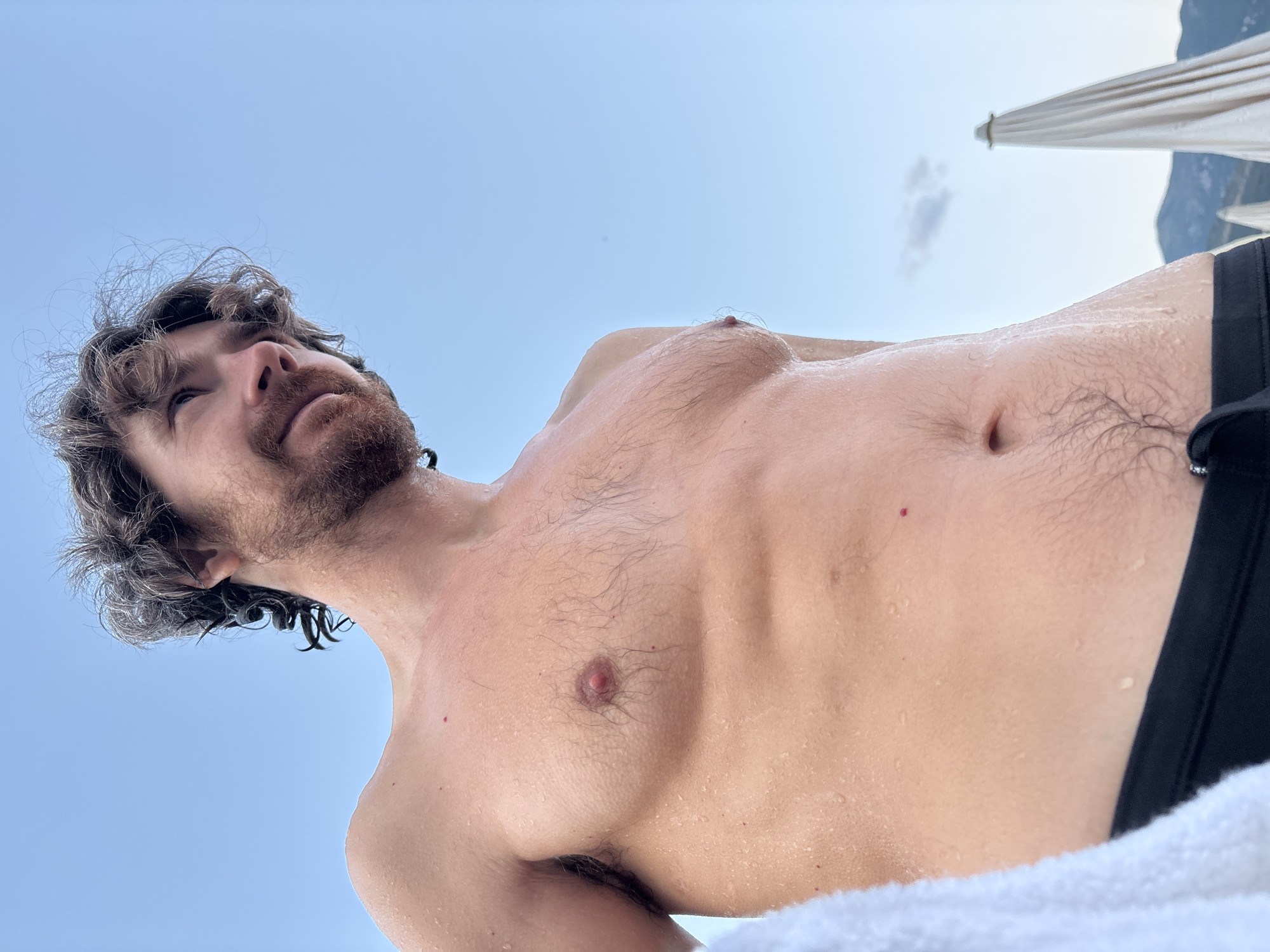 A man with wet, curly brown hair and a beard is looking up and to the right, with his chest and abdomen visible. He appears to be outdoors, possibly at a beach, with a clear blue sky in the background. There is a white towel on the left side of the image, and in the background, two closed beach umbrellas and mountains are visible. The man is wearing black swimming trunks. The overall impression is of a relaxed, post-swim moment. The location is identified as Boreti, Montenegro.