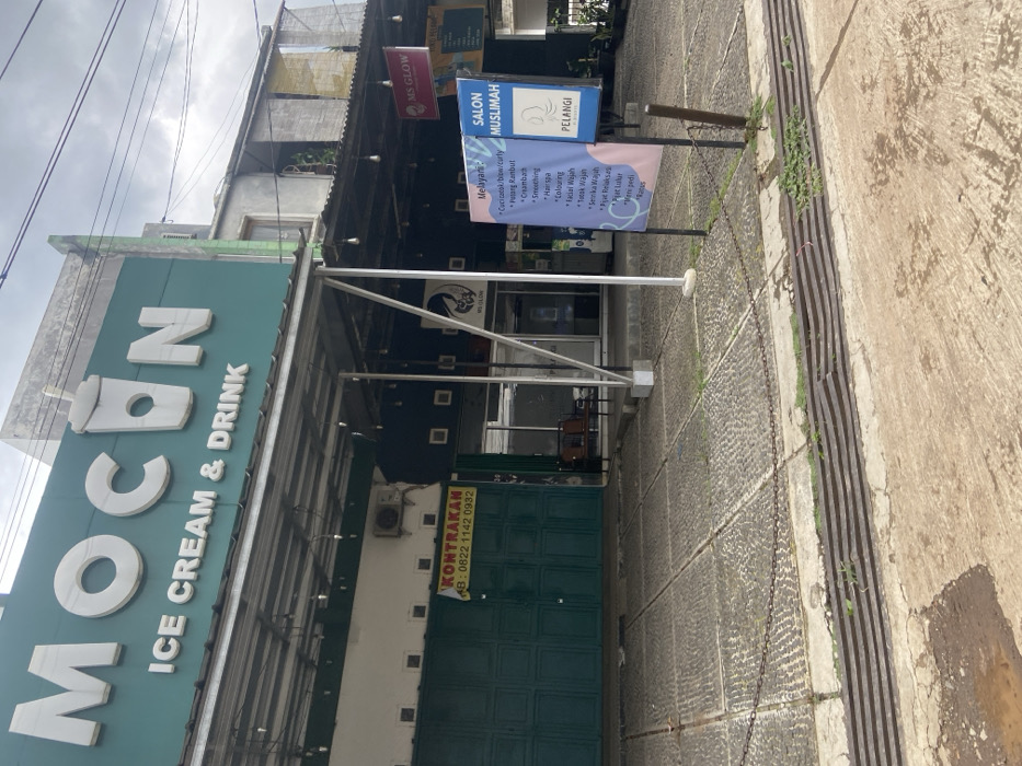 The image shows the exterior of two storefronts located at the coordinates -7.022, 107.676 in Baleendah, Indonesia. The storefront on the left is called "MOCON ICE CREAM & DRINK", with a green awning and a sign depicting a large cartoon glass of iced beverage. The storefront on the right is called "SALON MUSLIMAH" and appears to be a hair salon. The salon has a sign that lists various services, including "Creambath," "Smoothing," and "Hair Color." Both storefronts have chain-link fences extending from the sidewalk to the building. The ground in front of the stores appears to be a paved area consisting of small, irregularly shaped stones. The background of the image shows the sides of two buildings, partially obscured by the awning and the second floor of a taller building. The weather appears to be cloudy.