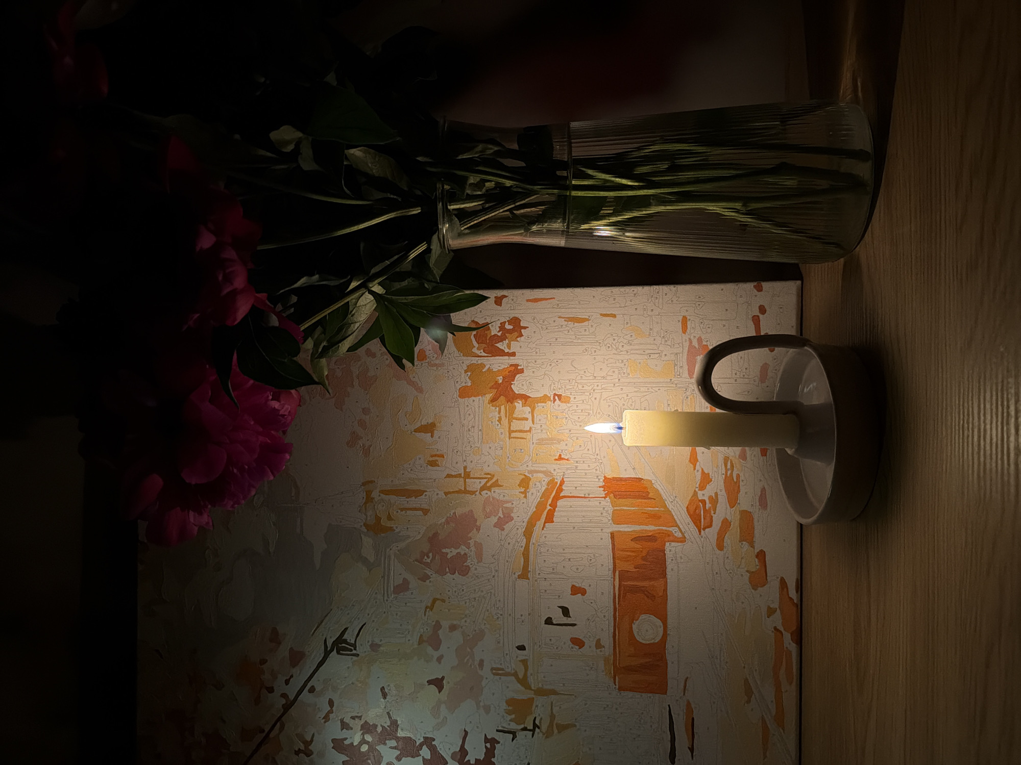 The image shows a dimly lit indoor scene with a paint-by-numbers canvas illuminated by a candle in a white holder. The canvas depicts an abstract cityscape in shades of orange, beige, and white. To the left of the canvas is a vase containing peony flowers. The environment appears to be a wooden surface. No people are visible. The city's location is unknown.