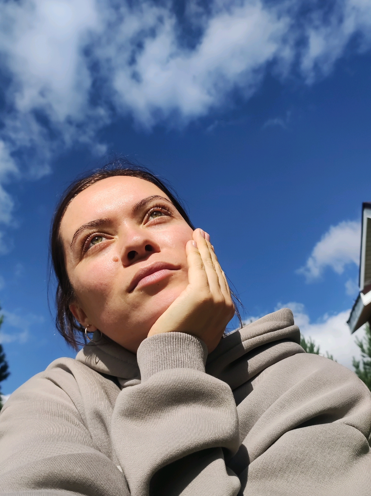 A young woman with brown hair and green eyes gazes upwards, her left hand resting on her cheek. She is wearing a light grey hooded sweatshirt. The background consists of a clear blue sky with scattered white clouds. The image appears to be taken outdoors, with the possibility of being in a suburban area due to a glimpse of a building and trees on the right side. The location of Фоки, Russia, is not ascertainable from the visual information provided in the image.