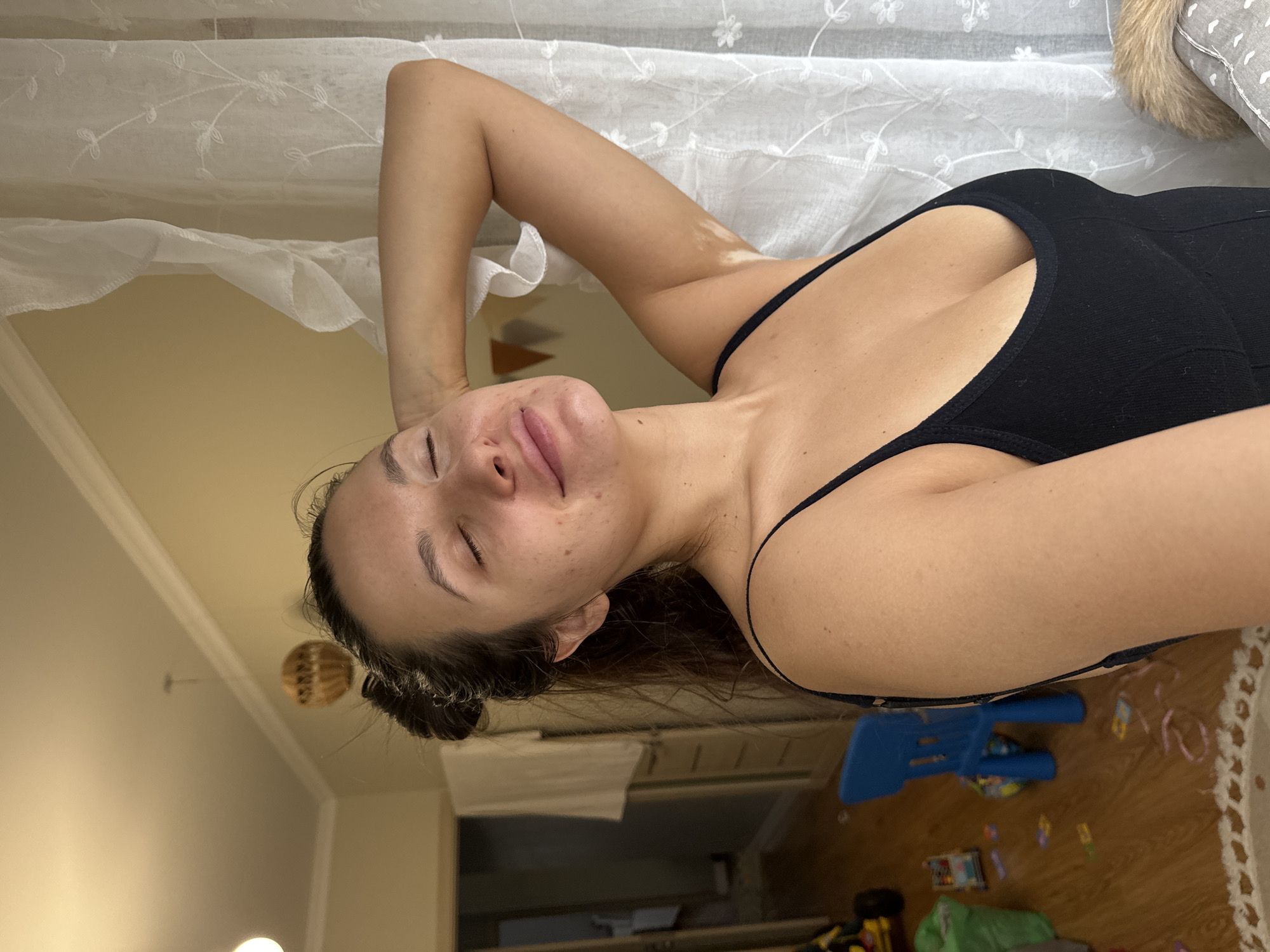 A medium shot captures a woman lying on a bed in a room. She is wearing a black tank top and her hair is pulled back. She is lying on her back, with her eyes closed and one arm raised above her head. The room has a white curtain, a wooden floor, and toys scattered around. The image was taken inside a residence, but its exact location in the city of Odintsovo, Russia, cannot be determined without additional information.