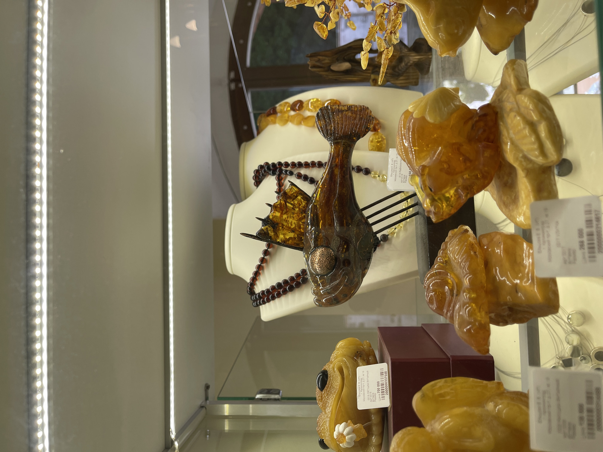 The image shows amber jewelry displayed in a glass showcase, likely inside a store in Kaliningrad, Russia. The jewelry includes a large fish pendant, a frog figurine, and several other amber pieces. Price tags with Russian text are visible, indicating the jewelry is for sale. One tag refers to "Ельцова ЕВ (ИП) Московский пр-т, д. 27, кв 19", suggesting a business owner located on Московский проспект (Moscow Avenue) in Kaliningrad at building number 27, apartment 19.