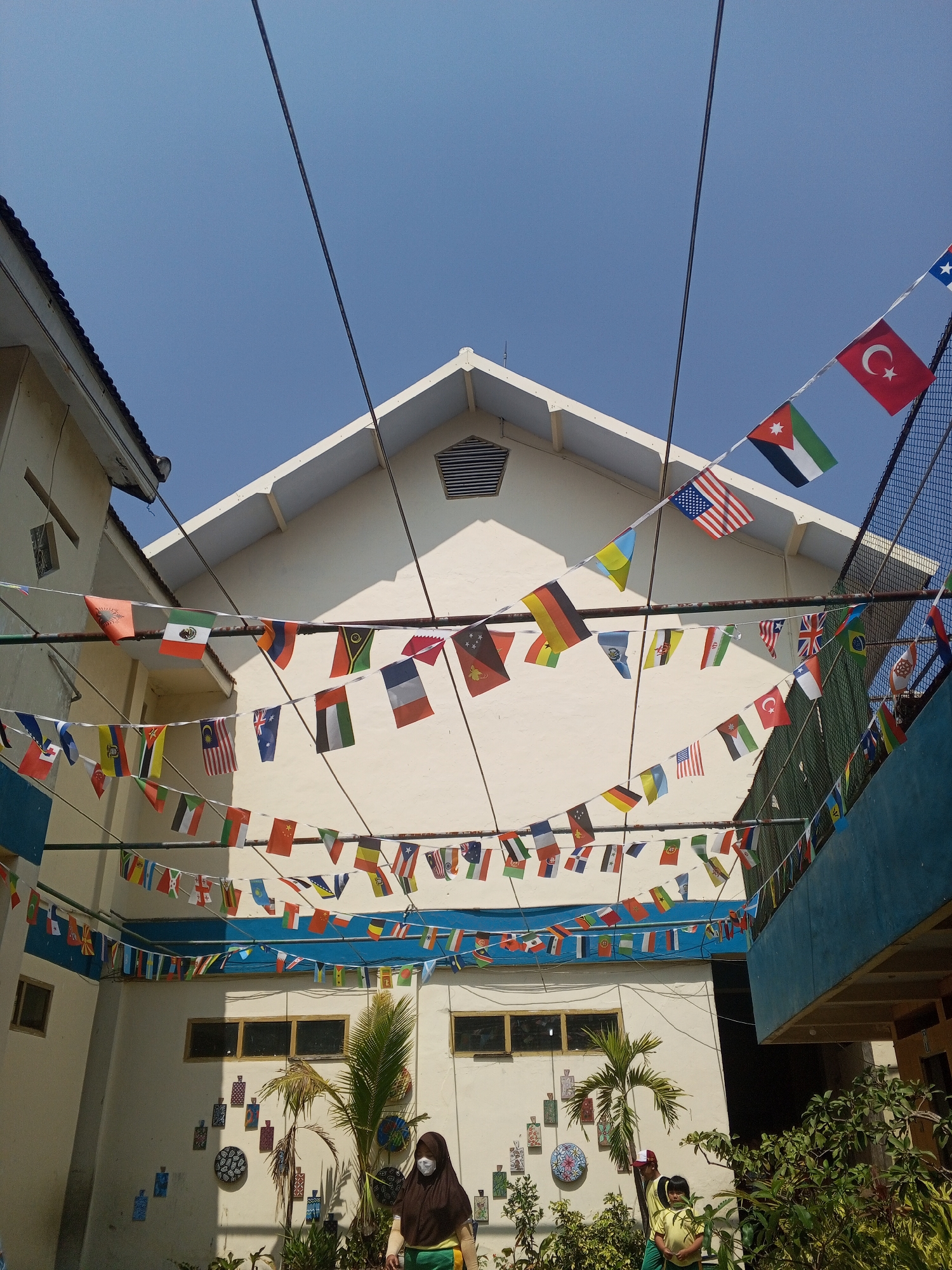 The image depicts the exterior of a building with a white facade, possibly a school, under a clear blue sky. The building has a pitched roof. Multiple strings of international flags are hung across the front of the building, creating a festive and multicultural atmosphere. The flags are small pennants, each representing a different country. Some of the visible flags include those of the USA, Turkey, Jordan, Australia, UAE, Germany, France, Brazil, and China. In the foreground, there are green plants and a palm tree. Two children in yellow shirts and green pants are visible, and an adult wearing a brown hijab and a white face mask is also present. The location is identified as Turen, Indonesia. The overall scene suggests an outdoor event or a permanent decoration celebrating diversity.