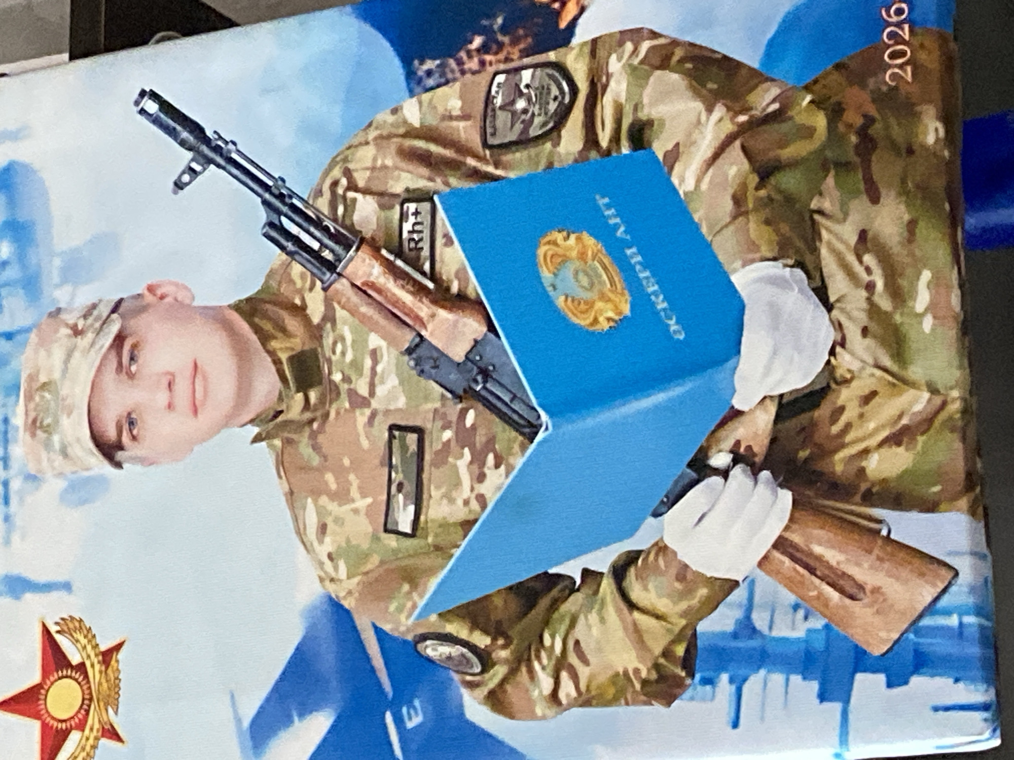 The image is a portrait of a young man in military uniform, holding a rifle and a blue folder. He is wearing a camouflage uniform, a cap, and white gloves. The rifle is an AK-style assault rifle, held across his chest and resting on the folder. The blue folder is open, revealing a gold emblem on the left page and Cyrillic text on the right page. The background of the portrait features a stylized depiction of fighter jets and a red star with a gold wreath, possibly symbolizing military achievement or national pride. On the lower right corner of the portrait, the year "2026" is visible. The text on the folder appears to be in Kazakh, with "ОСКЕРИ АНГ" clearly visible. The emblem on the left page of the folder is the national emblem of Kazakhstan. The uniform has several patches; one on the right sleeve depicts a circular emblem with an eagle, and another on the left chest area reads "Rh+". The overall presentation suggests a formal portrait, likely commemorating military service or a graduation from a military institution. The image is cropped from a larger display or print, as suggested by the slightly visible black border on the right side and the bottom. The portrait is situated in an indoor setting, given the lighting and the lack of any visible outdoor environmental features. The location specified as Shakhtinsk, Kazakhstan, is not visually depicted in the image itself, but the presence of the Kazakh national emblem and text strongly suggests a connection to Kazakhstan.