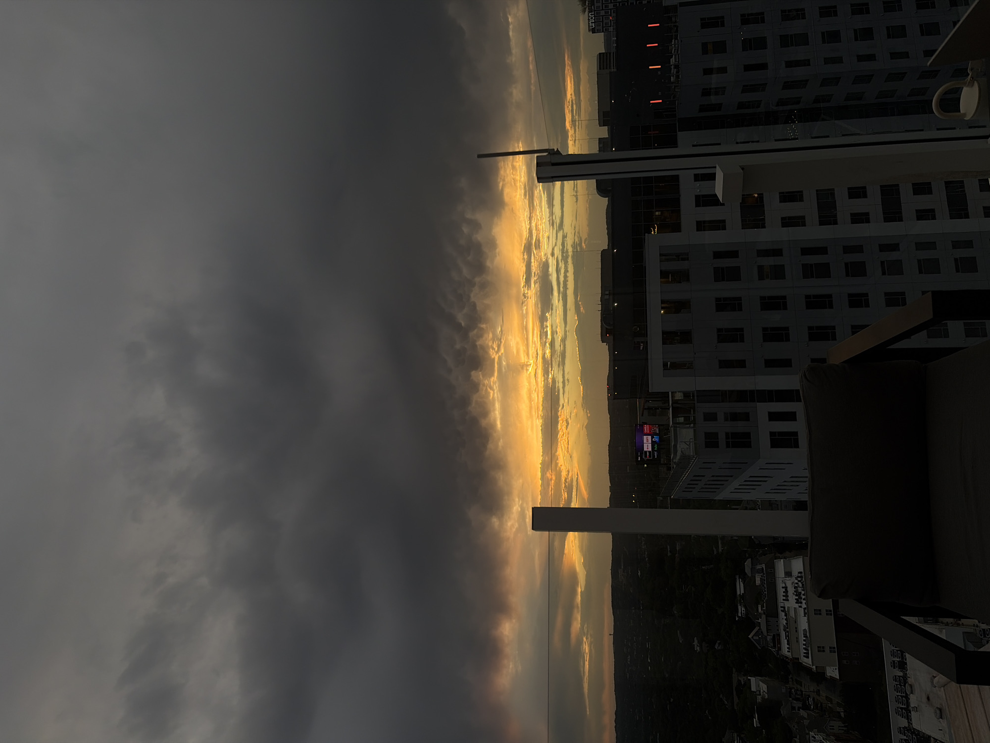 View from a high vantage point, likely a balcony or rooftop in the city of Austin, Texas, USA. The perspective is looking toward a cityscape, partially obscured by a dramatic sky with a strong contrast between dark storm clouds and a golden, sunlit horizon. Buildings of varying heights are visible, including residential and commercial structures. The sky is dominated by dark grey storm clouds on one side, and a bright, golden sunset or sunrise on the other. The built environment includes densely packed residential buildings, smaller structures along the ground, and green spaces. A portion of balcony furniture (chair or sofa) is visible in the foreground.