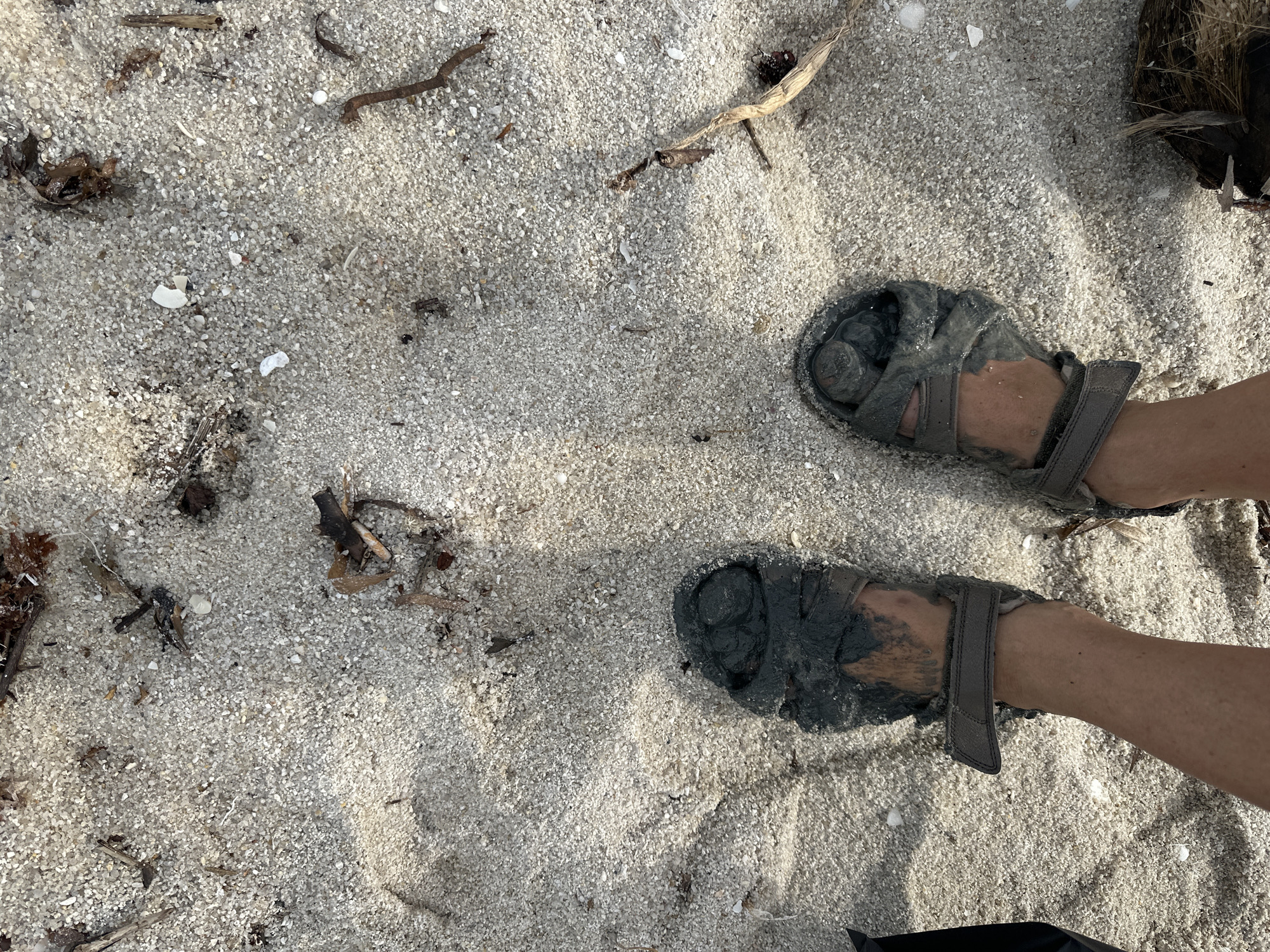 Two feet wearing muddy sandals are visible in the foreground, standing on a sandy beach. The sand is light-colored and appears to have small shells and debris scattered across it, including bits of dried seaweed and twigs. Shadows are cast on the sand, suggesting sunlight. The location is identified as a beach in Koh Phangan, Thailand.