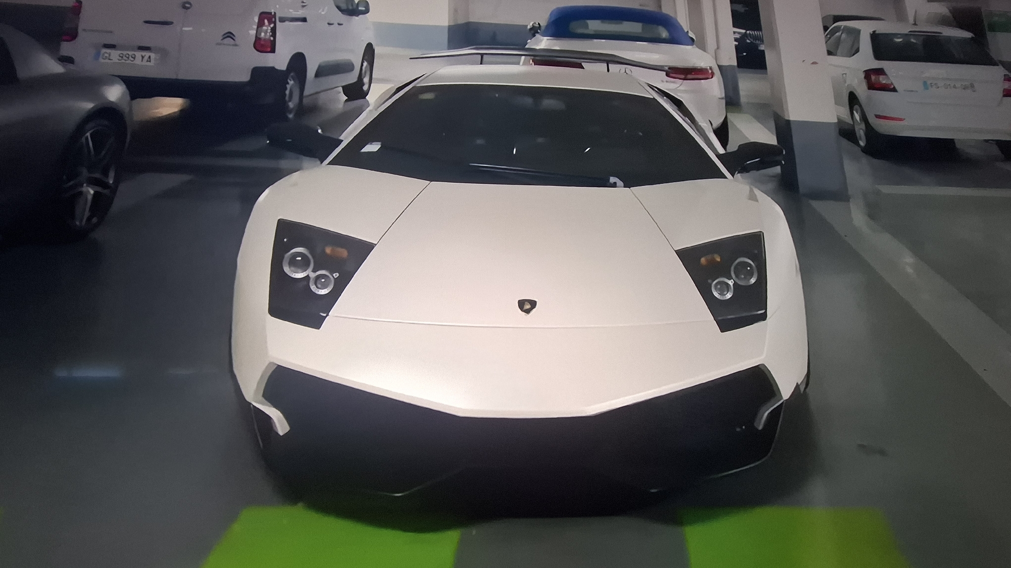 The image shows the front of a white Lamborghini parked in an underground parking garage. Other vehicles are visible in the background, including a white van with license plate "GL 999 YA," a light blue convertible, and a white car with license plate "FS-014-DR." The garage has light-colored walls and concrete flooring.
