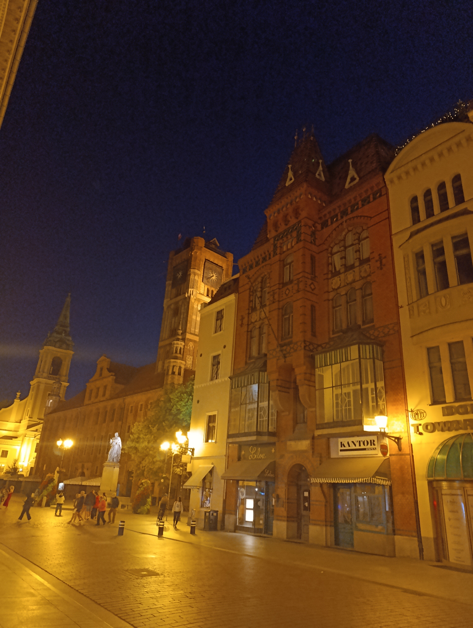 This is a low-light, nighttime photograph of a street scene in Toruń, Poland, likely in the Old Town area, given the architecture. The perspective is from a street level looking up at several buildings lining a cobblestone street.
On the left, a tall, green-spired building, possibly a church tower, is visible against the dark blue night sky. Adjacent to it, a large, red-brick building with multiple windows and ornate architectural details dominates the center and right side of the frame. This building appears to be several stories high and features a prominent clock tower with a sun-like face. In front of this building, another, lighter-colored structure with arched windows is partially visible.
The street itself is paved with cobblestones and is illuminated by warm, yellow streetlights, casting long shadows. Several streetlights are visible along the street.
A group of people, appearing as silhouettes due to the low light, are walking on the street. They are scattered, with some individuals walking alone and others in small groups. One person is wearing a red dress. In the middle of the street, a statue of a person on a pedestal is visible, surrounded by some greenery.
The building on the right is also illuminated by streetlights, highlighting its facade and windows. One building has a sign that reads "KANTOR" with "OPEN" below it. Another sign says "LILOU". The overall atmosphere is quiet and serene, characteristic of a European city at night.