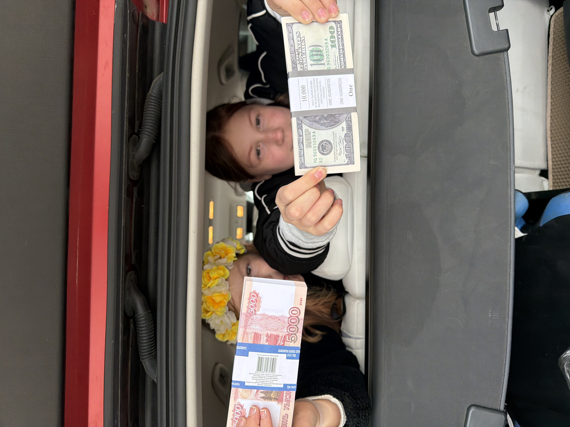 Two young girls are playfully posing for a photo while lying down in the rear cargo area of a vehicle. The image is taken from above, looking down at them.
The girl positioned higher in the frame has brown hair and a light smile as she holds a thick stack of novelty US 100-dollar bills. The wrapper on this stack explicitly states "SOUVENIR PRODUCTION" and includes text in Russian that translates to: "If the stated quantity is not enough in this package, it is possible that it is intended this way. Smile." It also indicates a value of "10.000" and "ONE HUNDREDS".
Below her, another girl with brown hair adorned with a yellow and white flower crown also smiles gently at the camera. She is holding a stack of novelty Russian 5000-ruble banknotes. The text on this stack also reads "СУВЕНИРНАЯ ПРОДУКЦИЯ" (Souvenir Production) and "БАНКНОТЫ ВАНКА ПРИ 2121 ГОД" (Banknotes of Vanka, year 2121), clearly indicating they are not genuine currency. The denomination "5000" and "ПЯТЬ ТЫСЯЧ" (Five Thousand) are also visible.
The setting appears to be the trunk or hatchback space of a car, with elements like red trim, black interior panels, and rubber seals visible. The girls are lying on a light-colored surface within the vehicle. Given the context of Russian ruble novelty money and the provided location, the scene takes place in Chaikovsky, Russia. The overall mood is lighthearted and playful, with the girls enjoying their photo opportunity with the fake money.